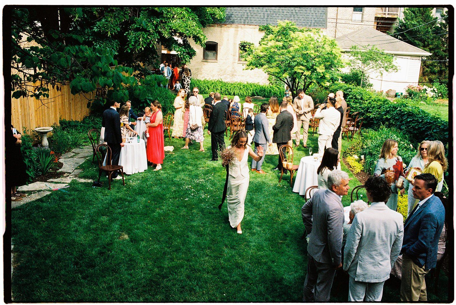 People gathered in a backyard garden for a social event, with several tables, chairs, and lush greenery.