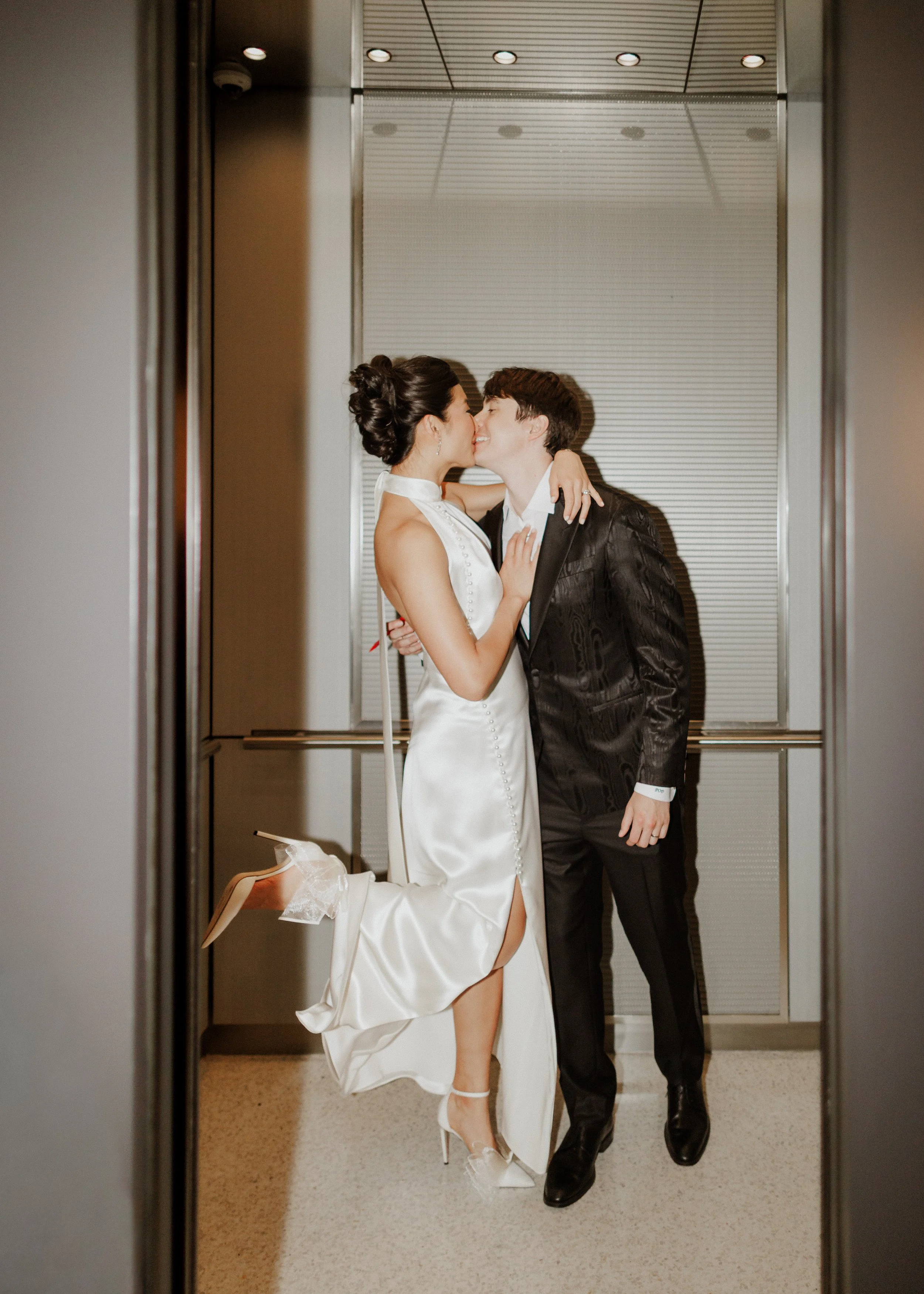 A woman in a white wedding dress and high heels sharing a kiss with a man in a black suit inside an elevator.