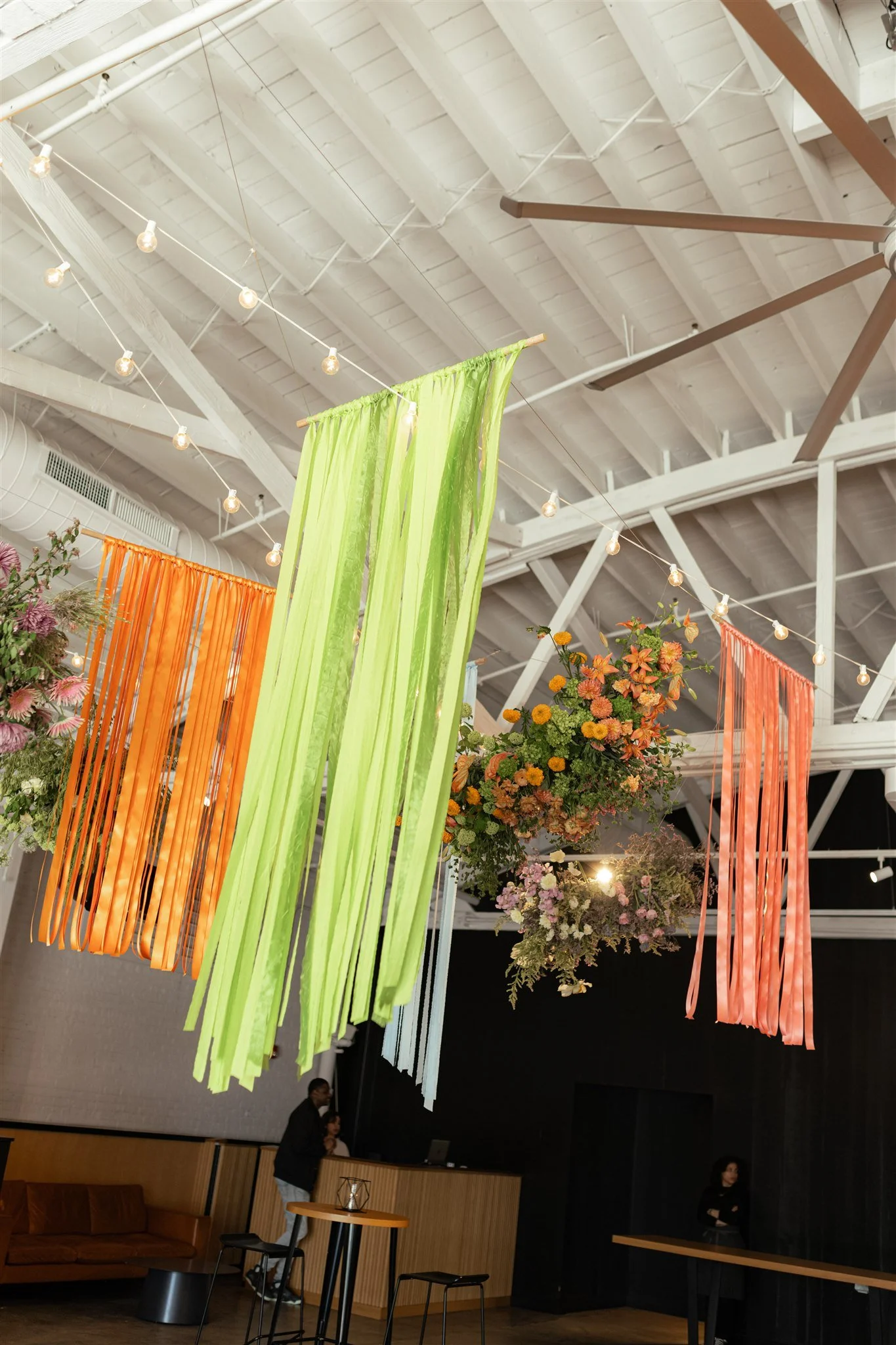 Interior of a modern café or lounge area with hanging colorful fabric strips in green, orange, and light blue, decorated with string lights and floral arrangements on a high ceiling with exposed beams.