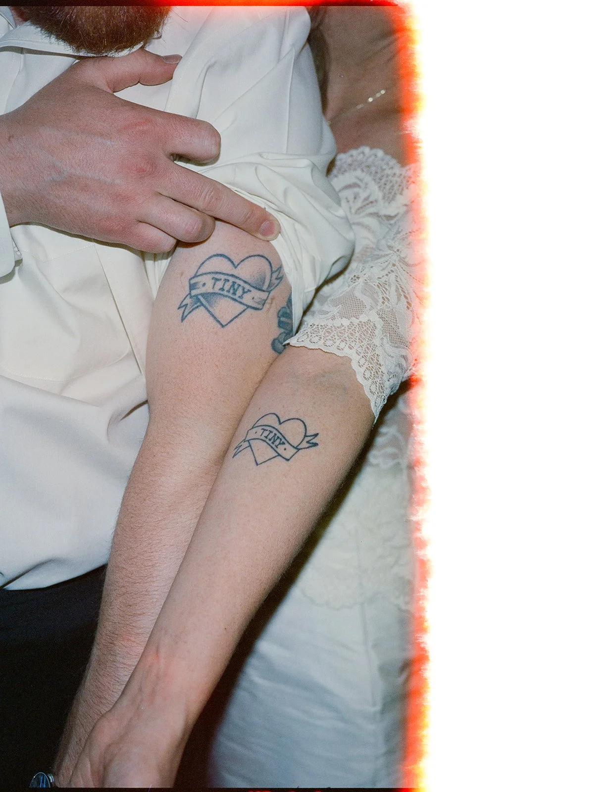 Person holding another person's arm, revealing matching tattoos of a heart with a banner that reads 'TINY' on the upper arm.