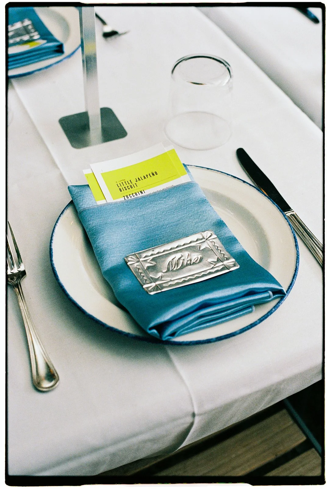 A place setting includes a white plate with a blue rim, a folded blue napkin with a silver nameplate, and a yellow menu card. Silverware is placed on each side of the plate. A clear drinking glass and a partially visible small stand are on the table,