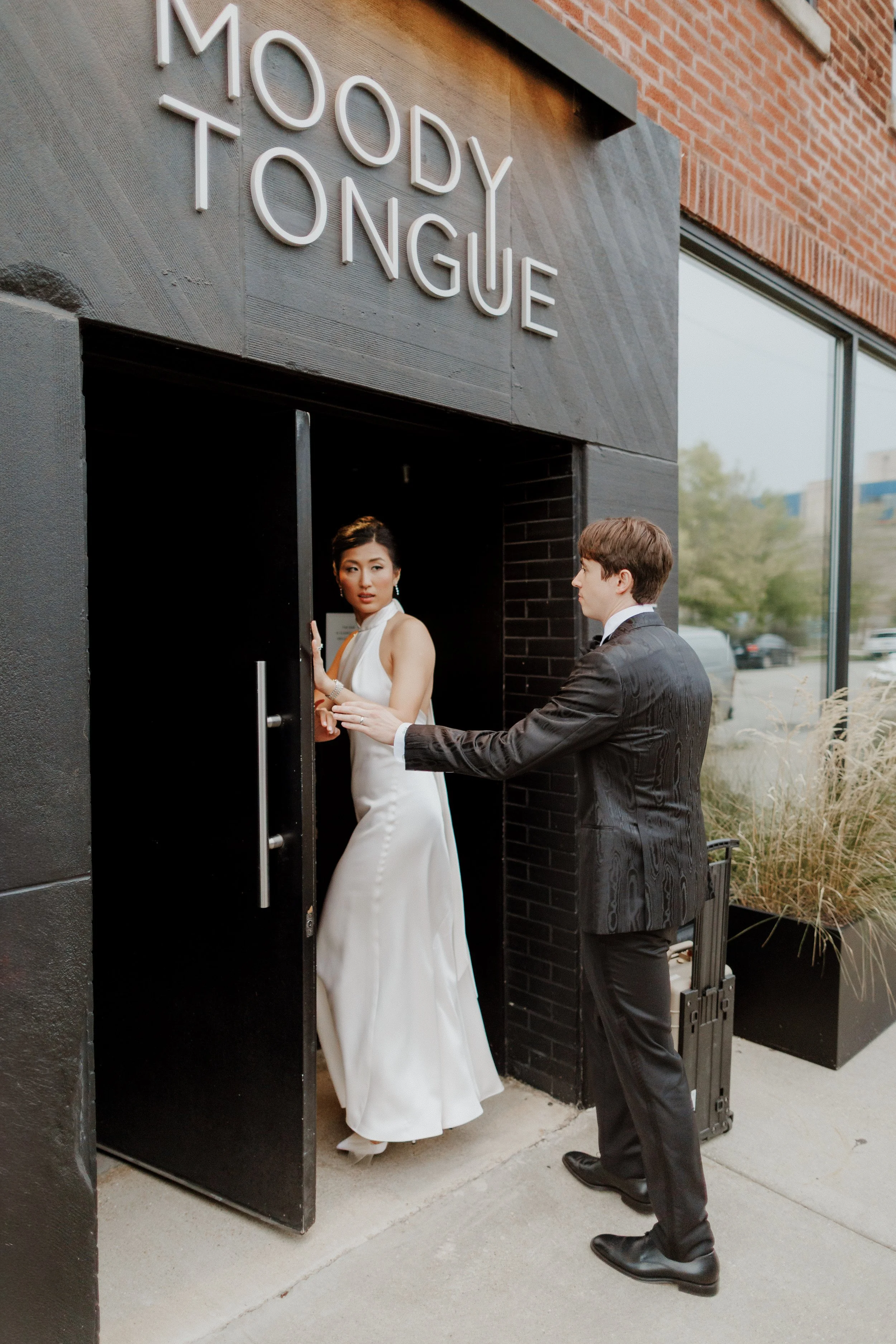 A bride in a white gown with a high slit and a groom in a black suit standing outside the entrance of a building with a sign that reads 'MODOBY TONGUE'. The bride is entering the building, and the groom is holding the door open for her.