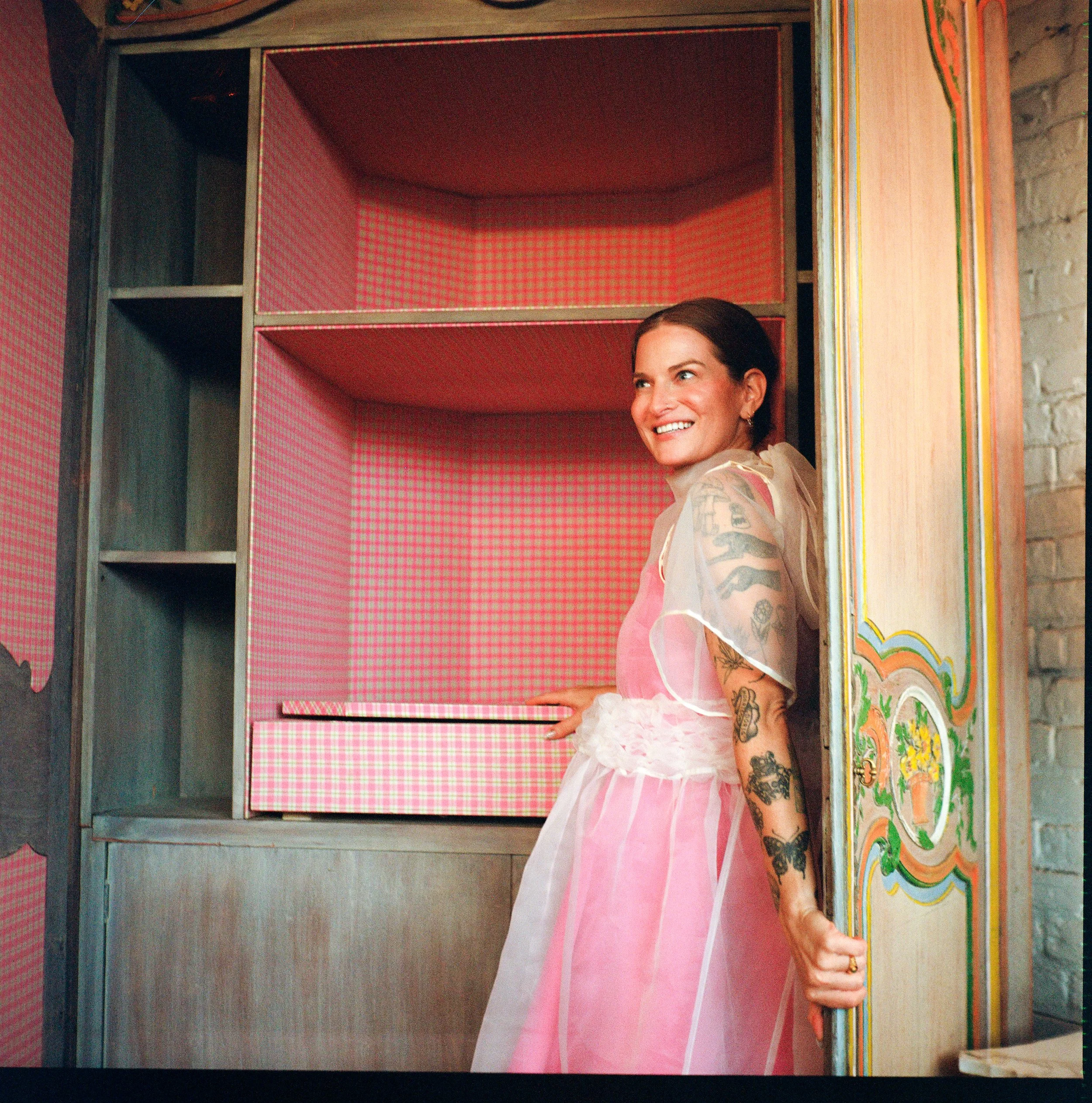 A smiling woman with dark hair and tattoos on her arms wearing a pink dress is standing inside a partially open colorful wooden structure with pink checked fabric lining.