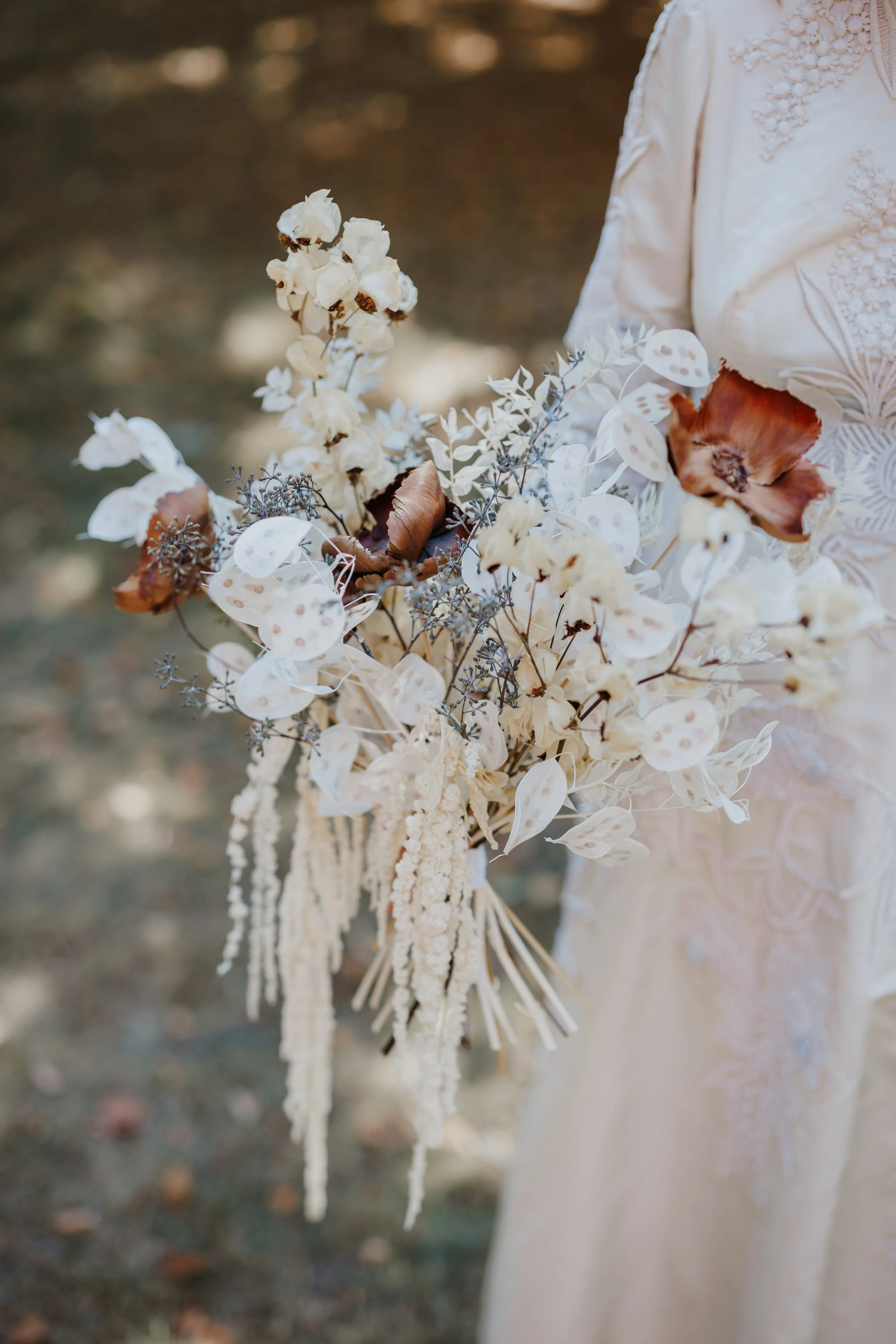 A person holding a bouquet of dried flowers, including cotton, matte leaves, and brown and cream-colored blooms, while wearing a cream dress with lace and embroidery details.