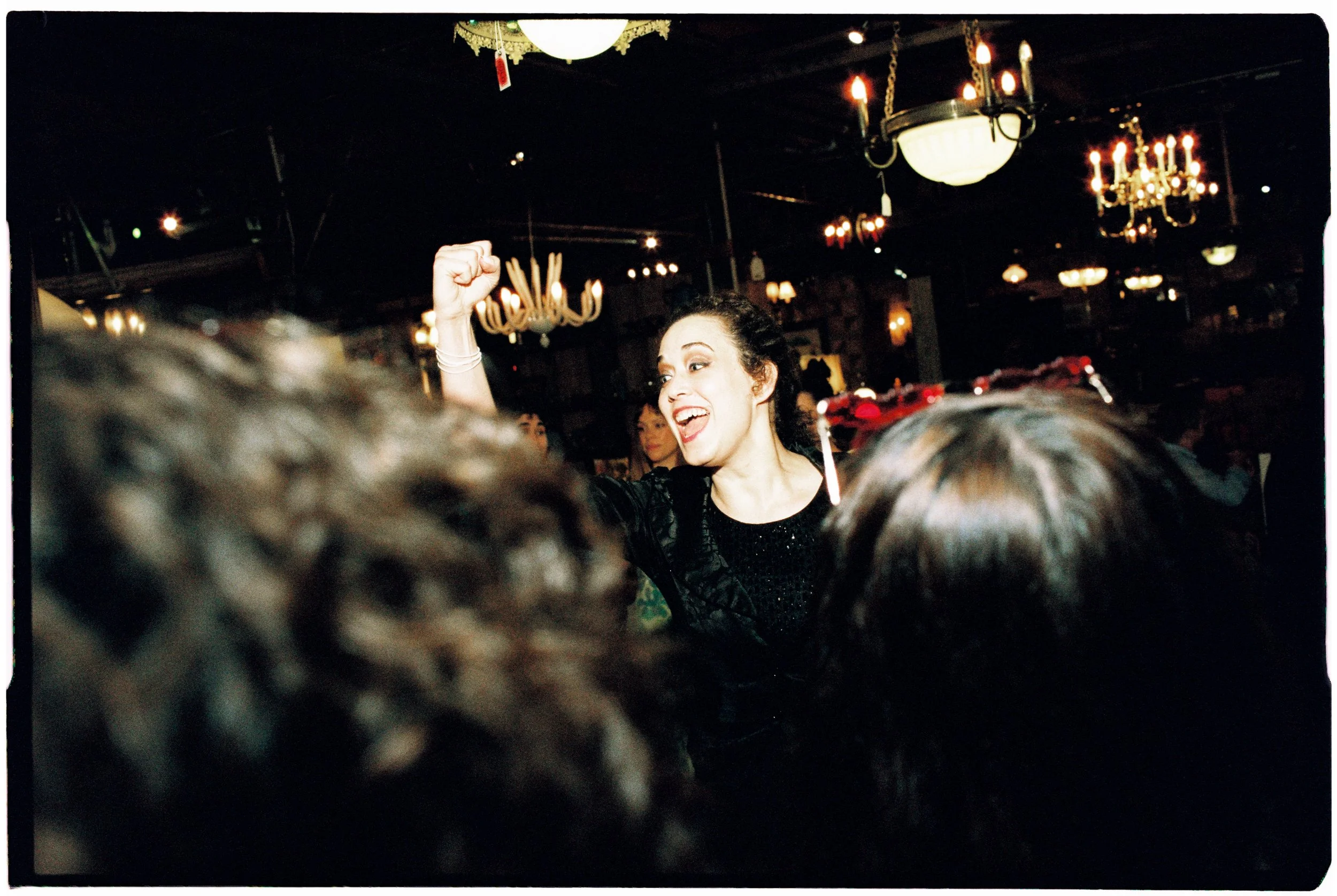 A woman with dark curly hair, wearing a black dress, smiling and raising her fist in celebration at a crowded indoor event with chandeliers hanging from the ceiling.