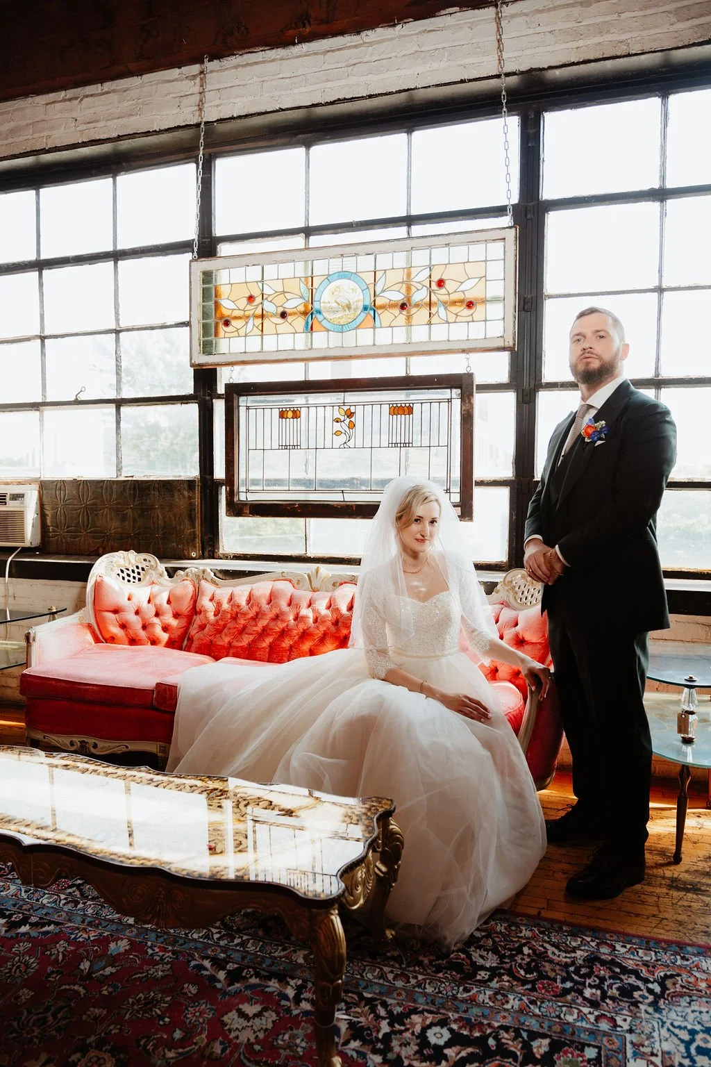 Bride in white gown sitting on a vintage red sofa with groom standing beside her in a black suit, inside an industrial-style room with large windows, stained glass decorations, wooden floor, and vintage furniture.