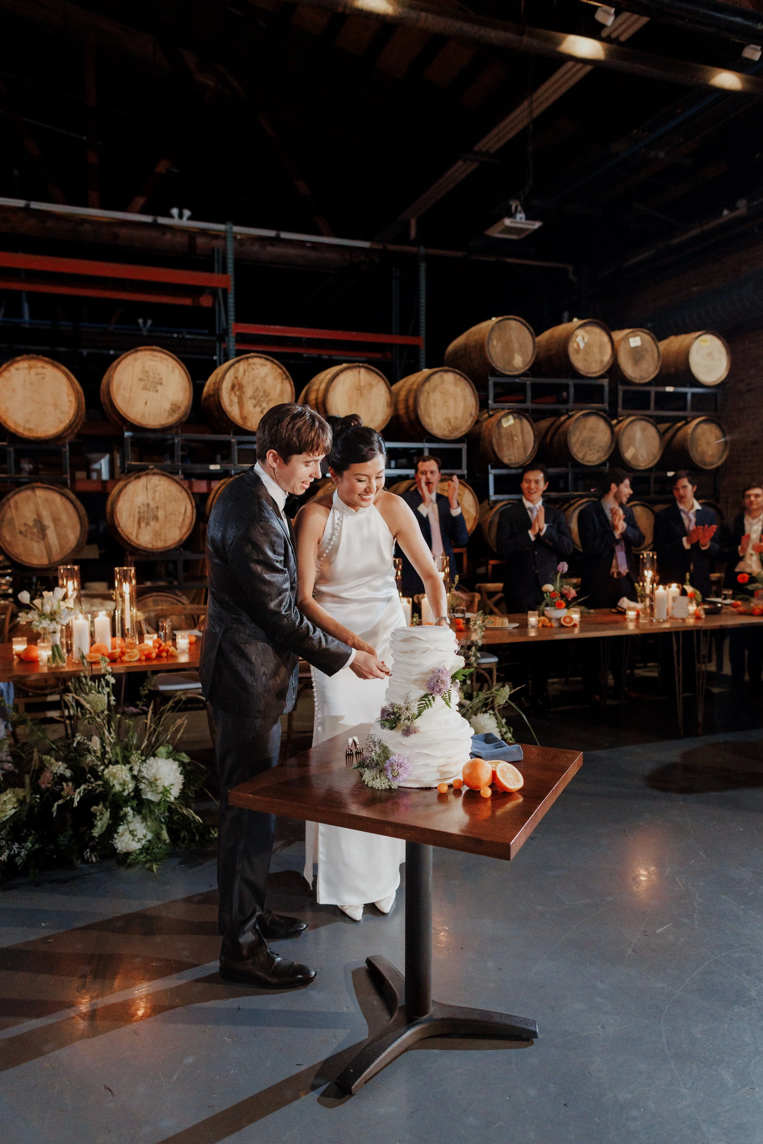 A newlywed couple cutting a wedding cake at a reception in a rustic venue with wine barrels in the background. Guests are clapping and celebrating around them.