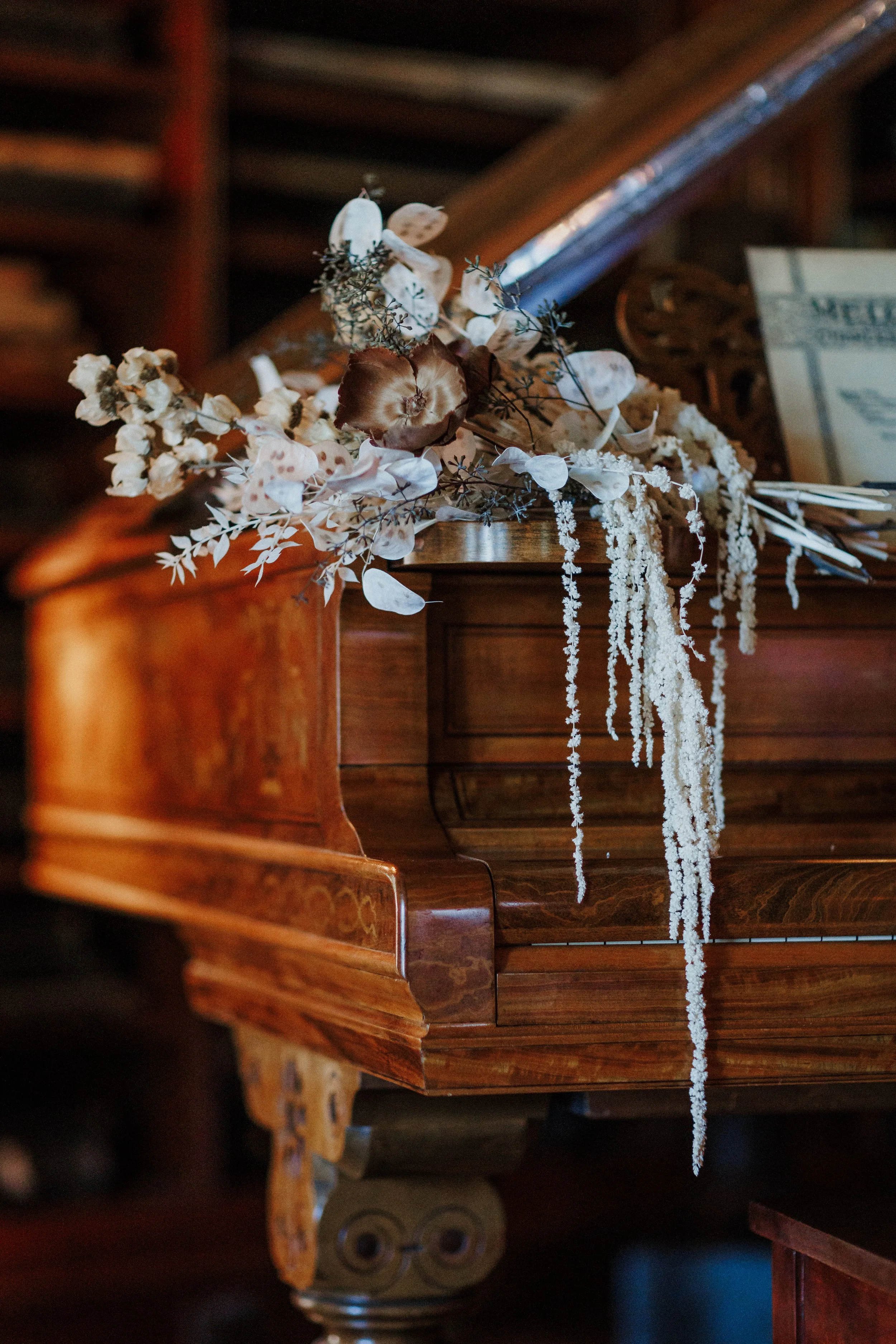 A close-up of vintage wooden furniture with a floral arrangement on top, featuring white and brown dried flowers, and a portion of a decorative column and a newspaper in the background.