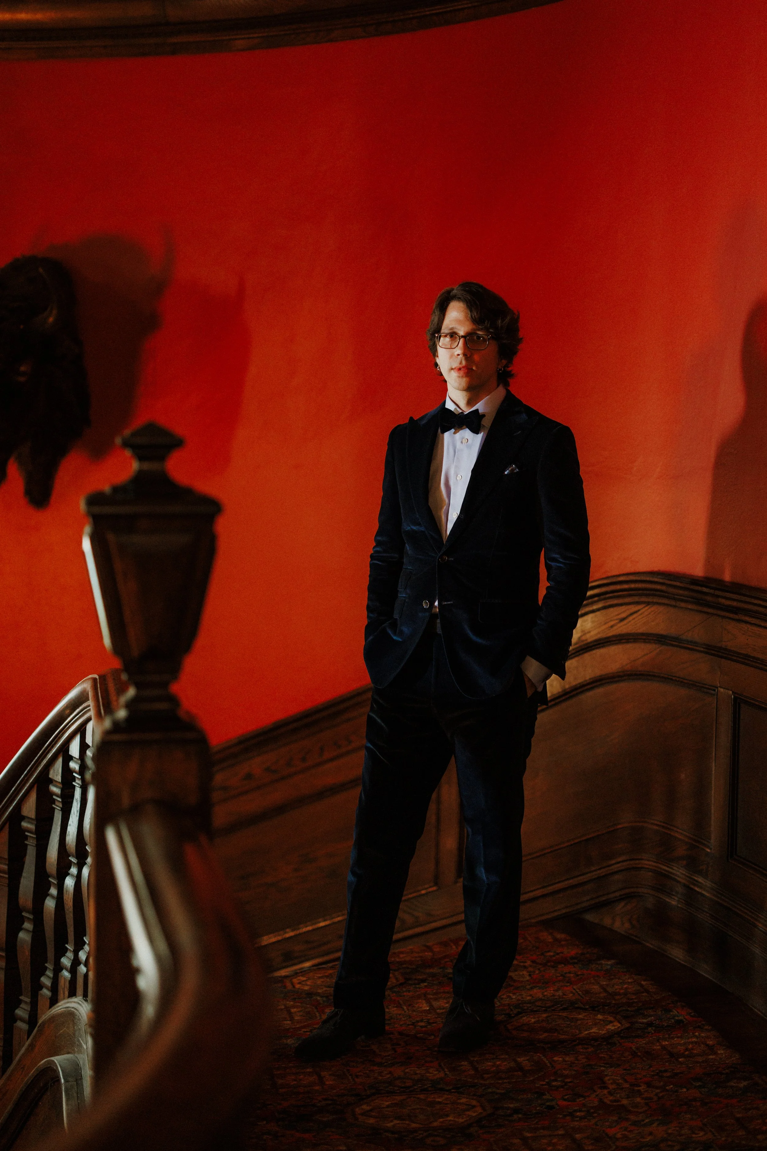 A man wearing glasses in formal attire with a tuxedo and bow tie, standing on a staircase with wood paneling and a red wall in the background.