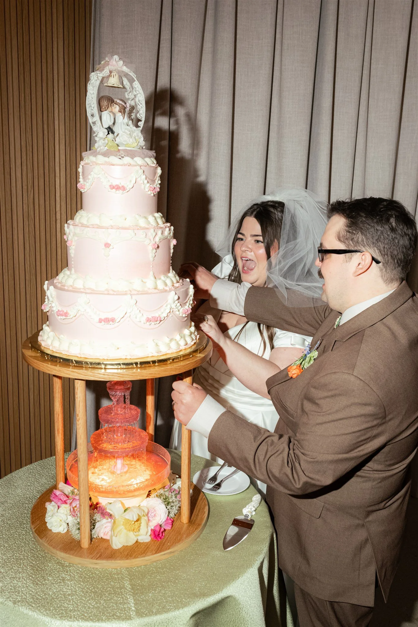 A bride and groom cut a pink multi-tiered wedding cake with a figurine topper of a kissing couple. The cake features floral decorations and is displayed on a wooden stand with flowers around its base. The bride is wearing a white wedding dress and ve