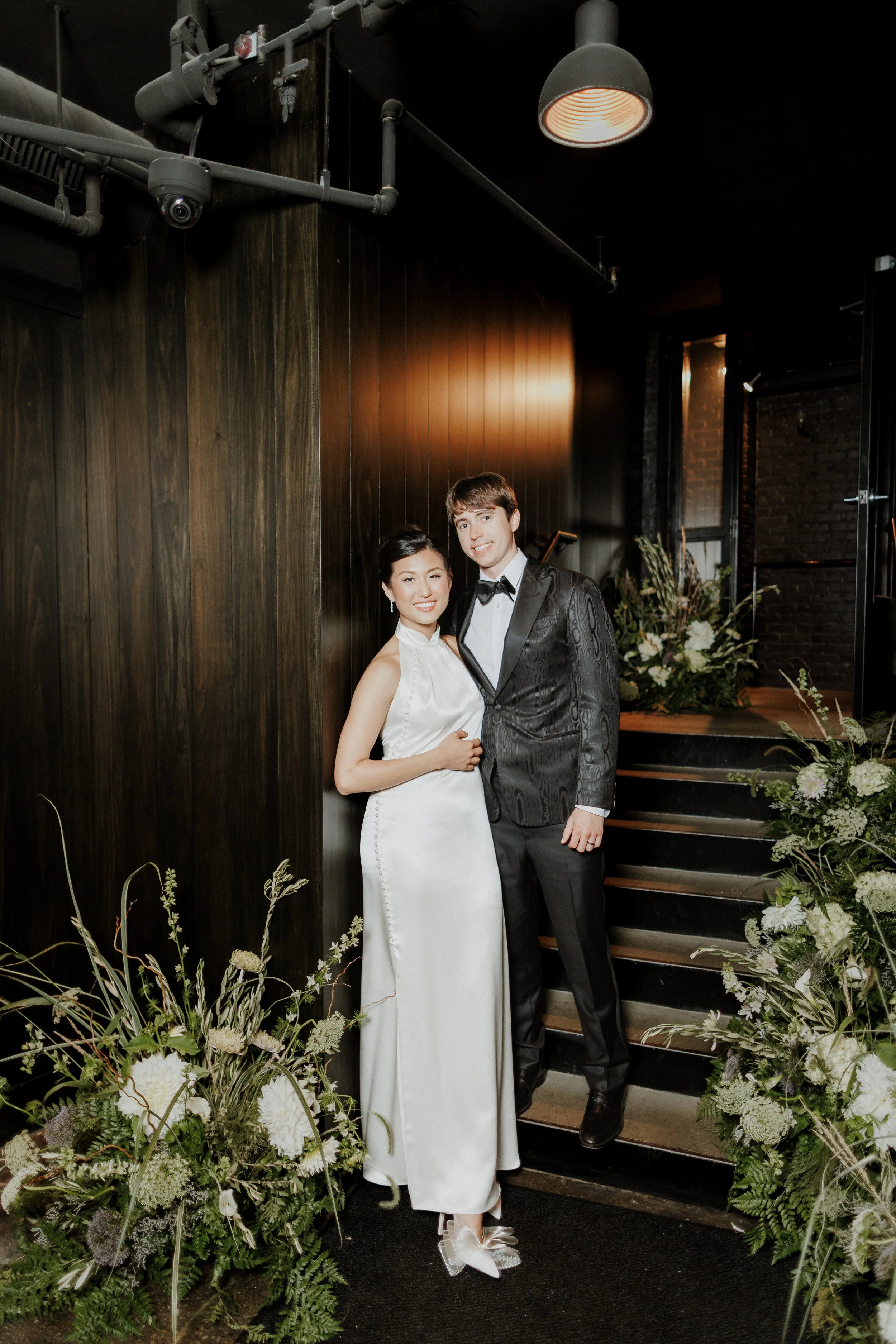 A bride and groom standing close together on a staircase, surrounded by floral arrangements, inside a dimly lit venue. The bride is wearing a sleeveless white gown with buttons down the back and bow shoes, while the groom is in a black tuxedo with a 