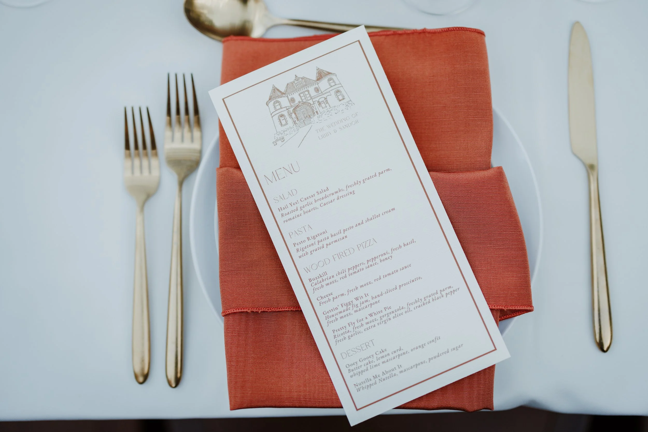 A wedding menu placed on a red napkin with gold utensils on a table. The menu features a drawing of a large house or mansion and lists various food items, including salads, pasta, pizza, and desserts.