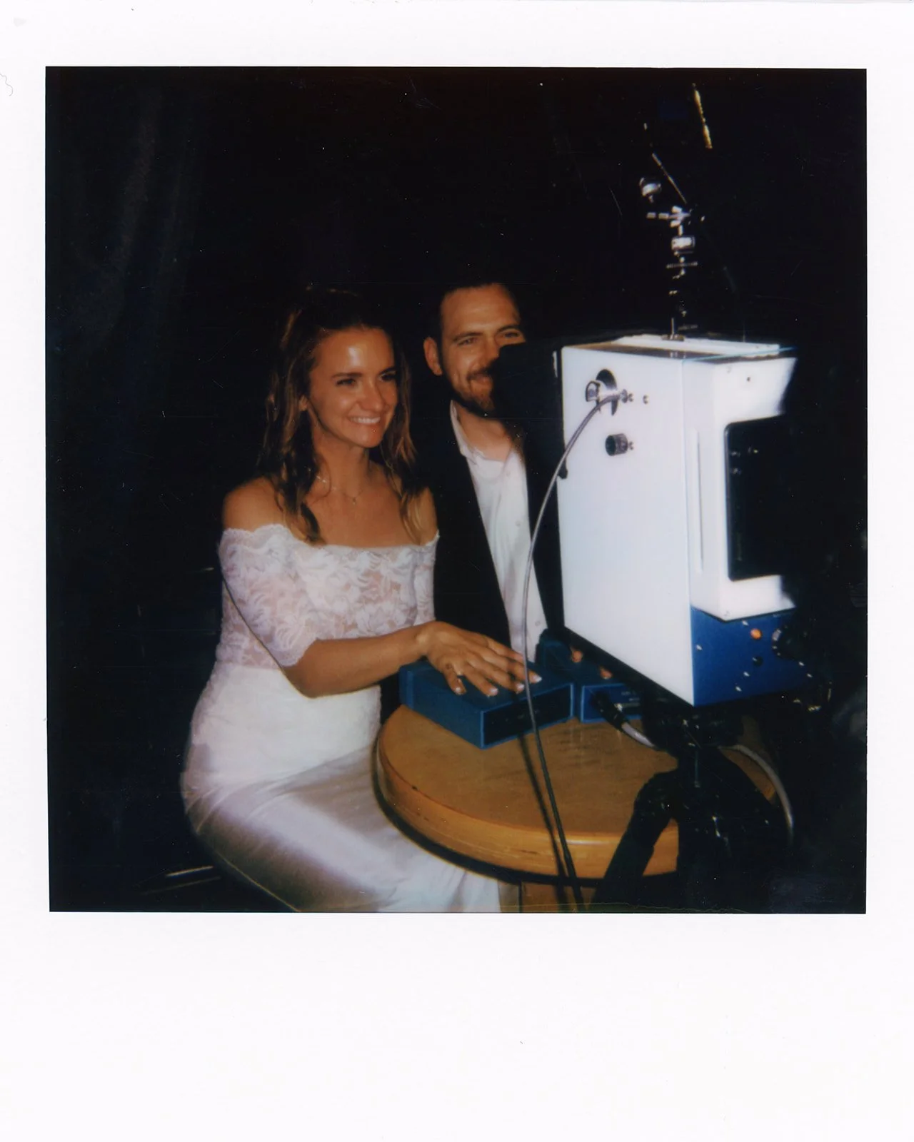 A woman in an off-the-shoulder lace white dress and a man in a suit standing behind a vintage television on a round wooden table, smiling for the camera.