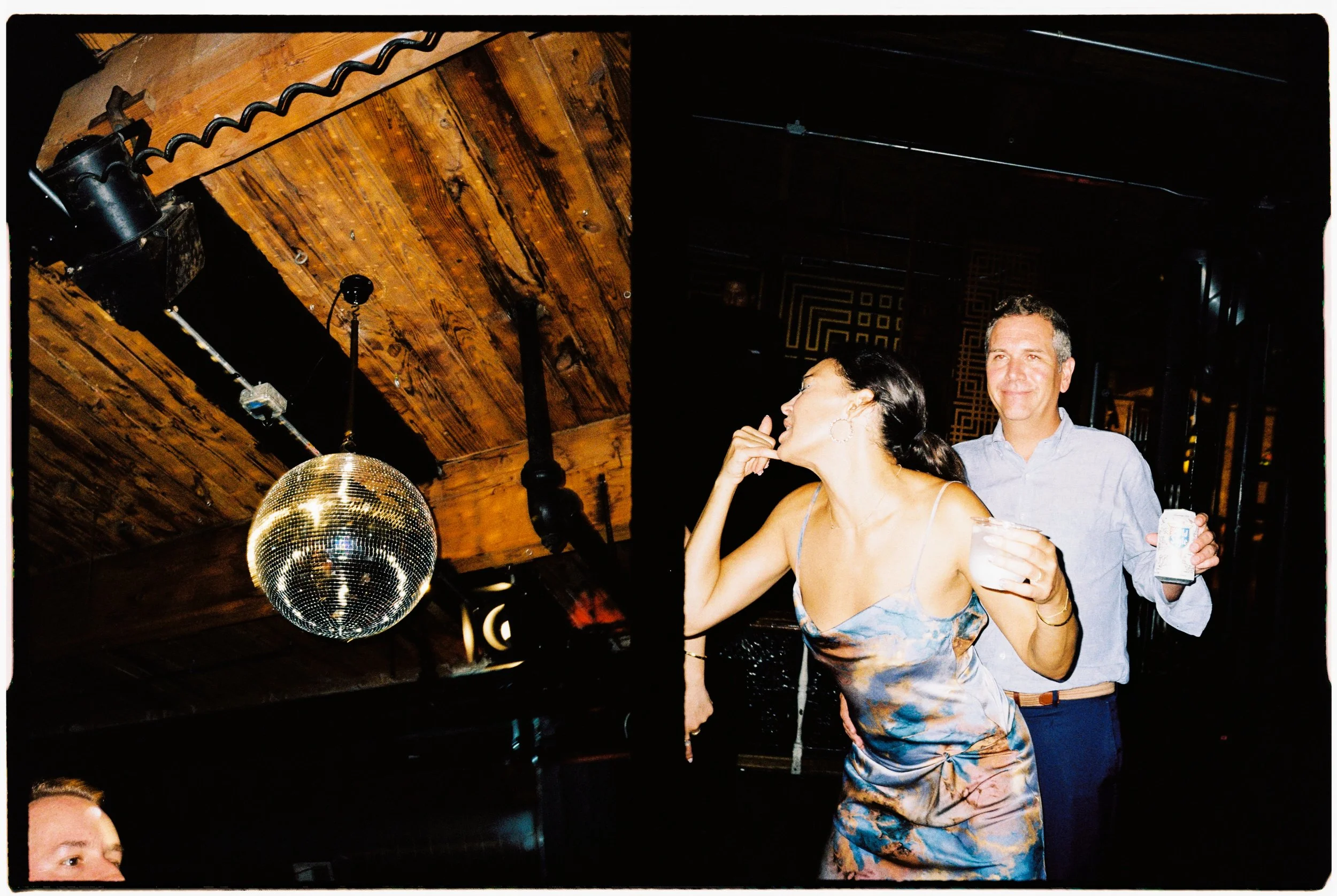 A woman with dark hair and hoop earrings wearing a blue and orange floral dress, talking and smiling, with a man standing behind her wearing a light blue shirt, holding a drink in a glass, in a dimly lit venue with wooden ceiling and a disco ball han