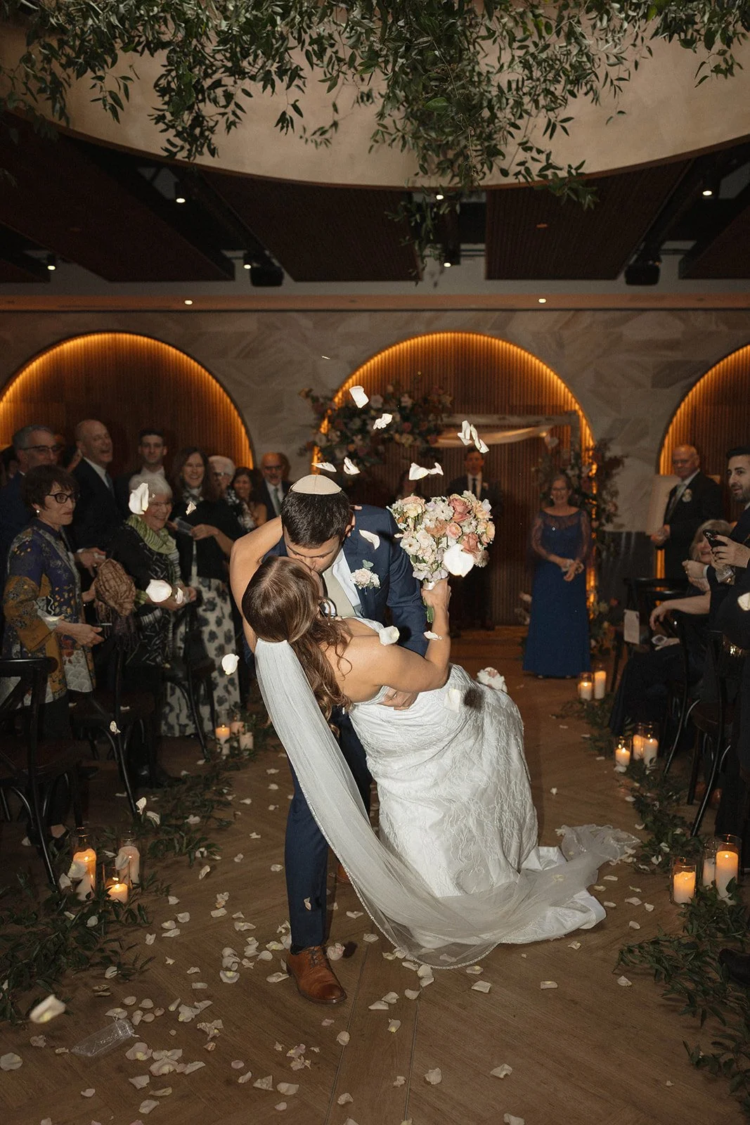 A wedding couple dancing closely during their wedding reception, surrounded by guests, candles, and flower petals.