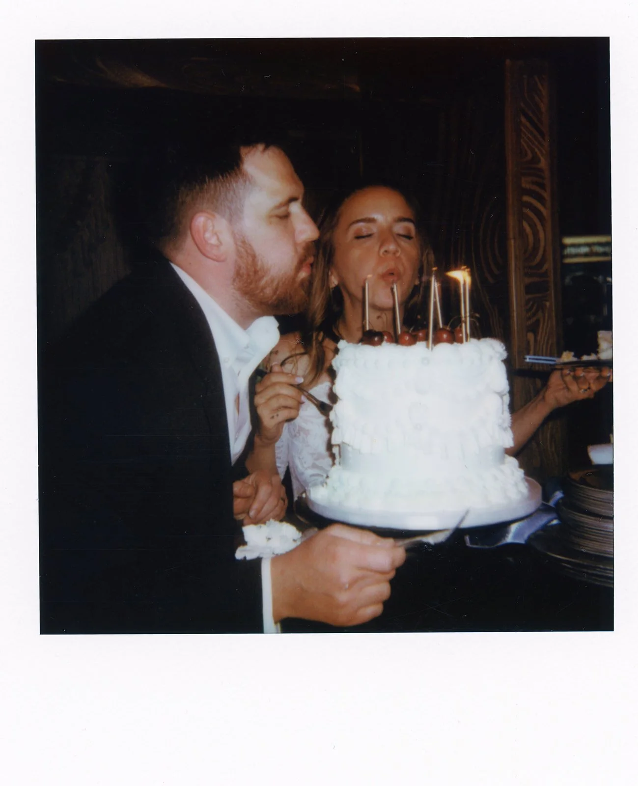 A newlywed couple dressed in formal attire, with the man in a tuxedo and the woman in a wedding dress, blowing out candles on a wedding cake at their celebration.