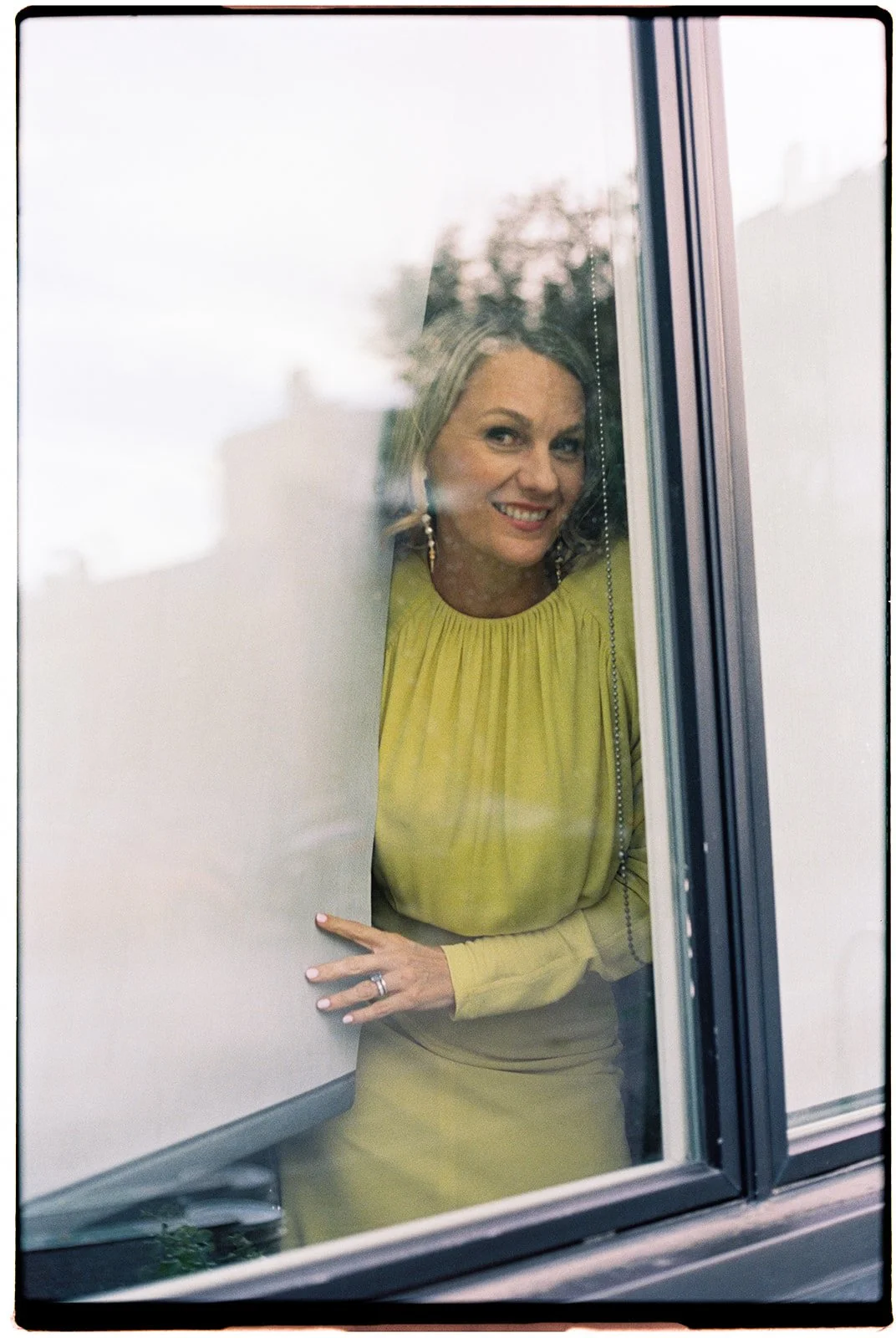 A woman in a yellow dress smiling and looking through a window from the outside.