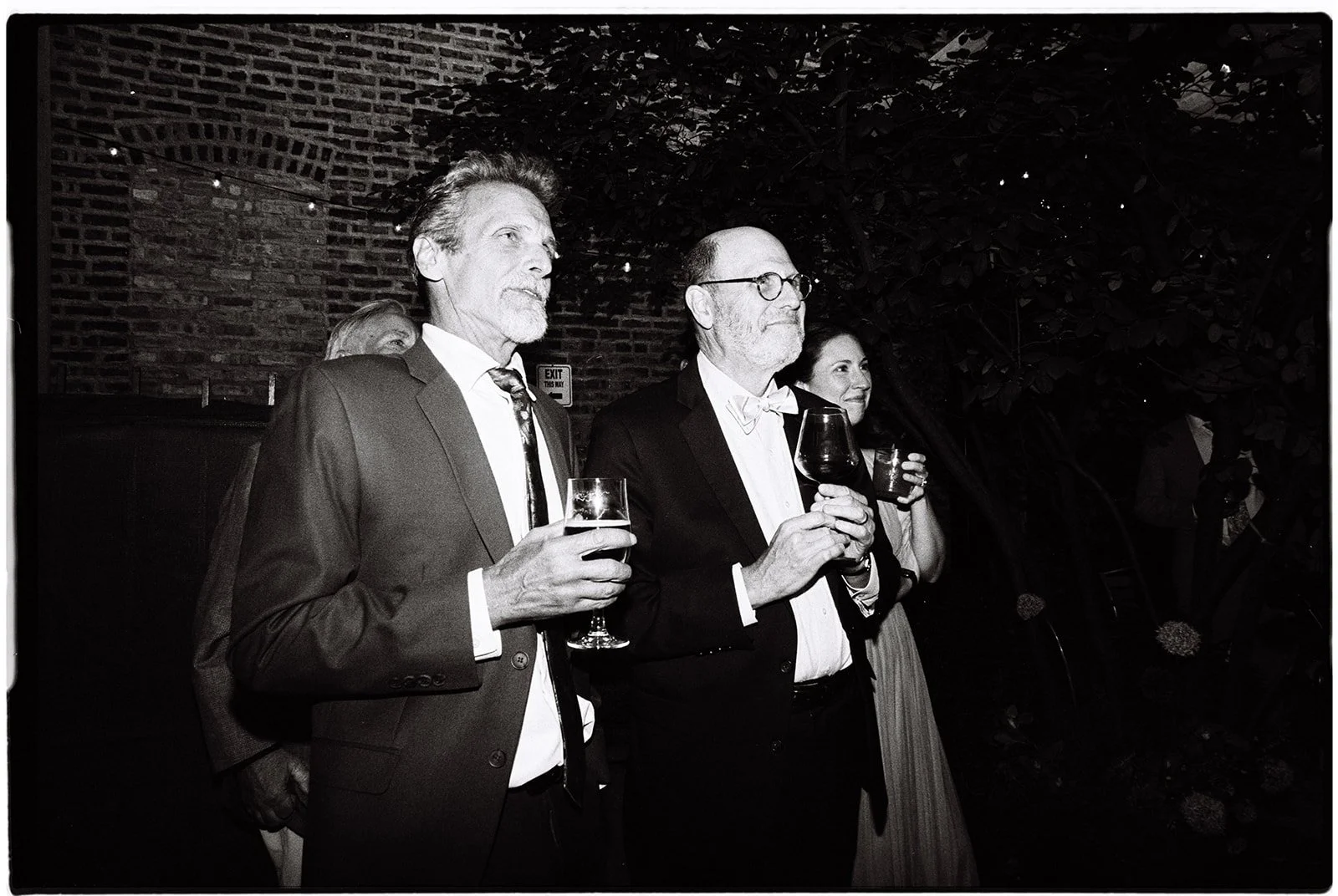 Three people in formal attire holding glasses of wine, standing in a dimly lit, outdoor setting with a brick wall and foliage in the background.