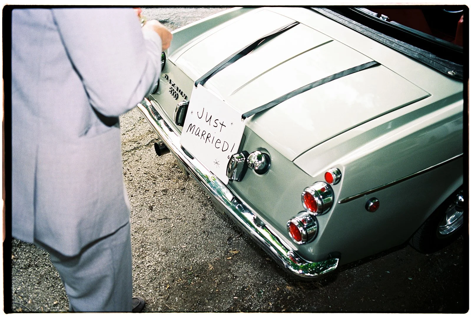 A silver vintage car with a sign on the back that reads 'Just Married' in black marker, and a person standing nearby in a light gray suit.