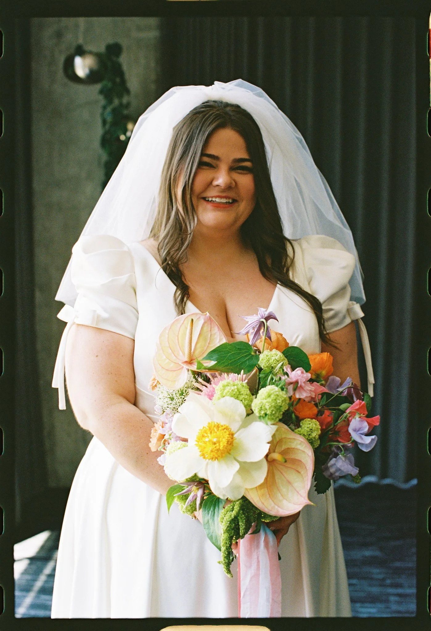 A woman in a white dress and veil holding a colorful bouquet of flowers, smiling at a special occasion or event.