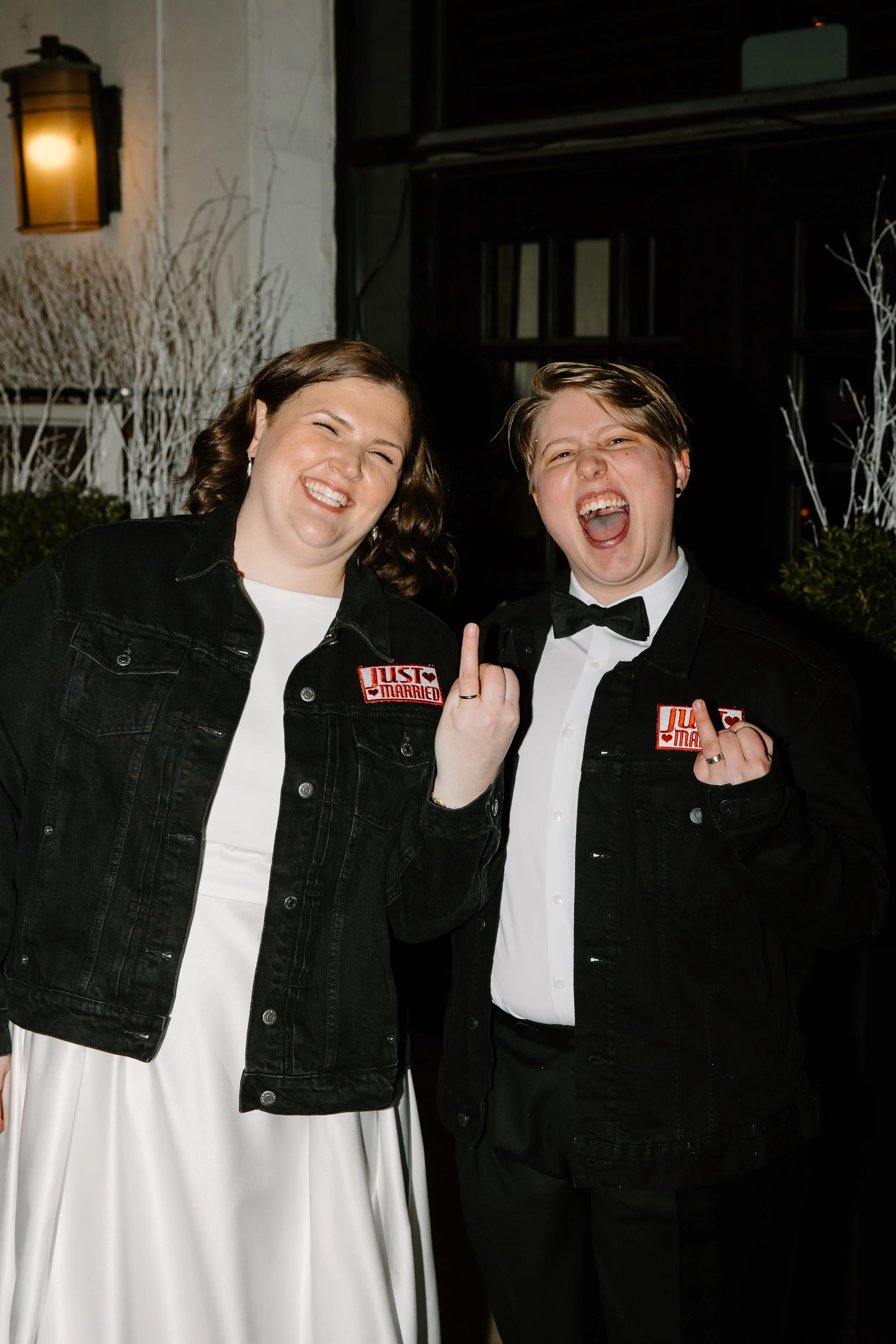 Two people smiling and making a rebellious gesture while wearing black jackets with 'JUST MARRIED' patches, standing outdoors at night.