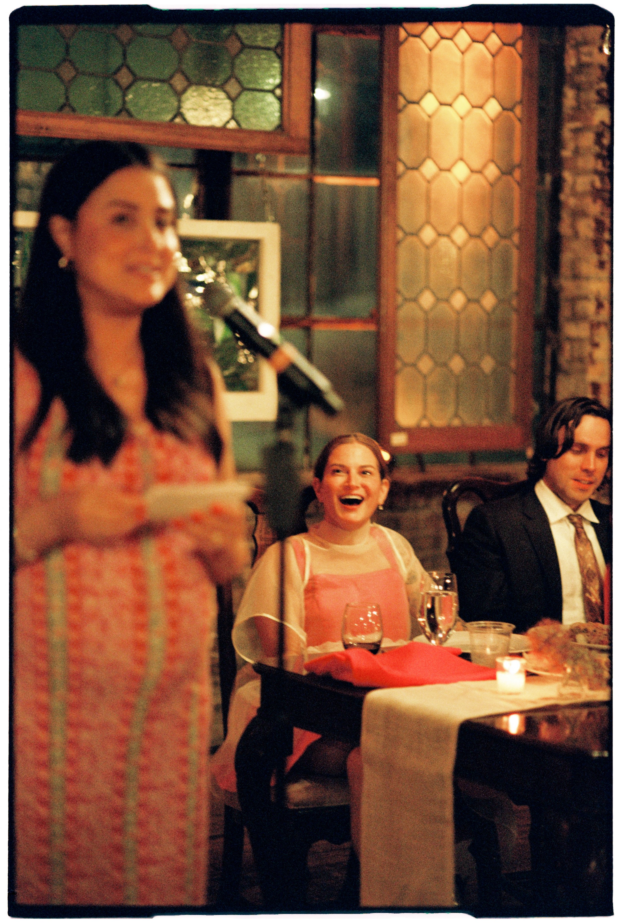 A woman speaking into a microphone at a restaurant or banquet setting, with three other people seated at a table, including a woman laughing and two men, one of whom is dressed in a suit and tie.