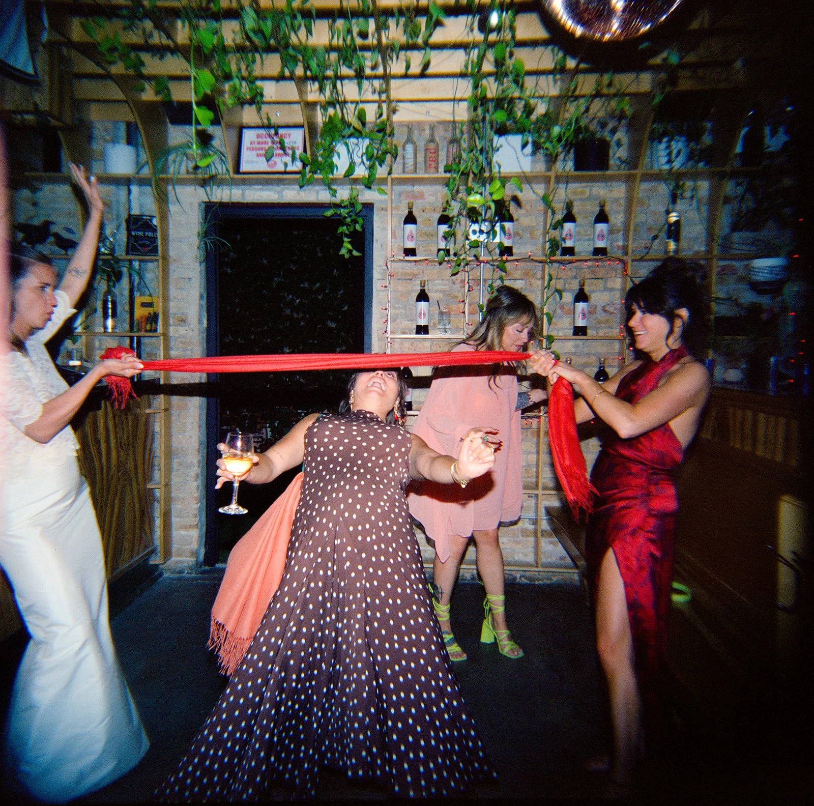 Four women celebrating at a party or bar, one holding a glass of wine, and others engaging in a playful activity with a red cloth, in a brick-walled setting with bottles and hanging plants.