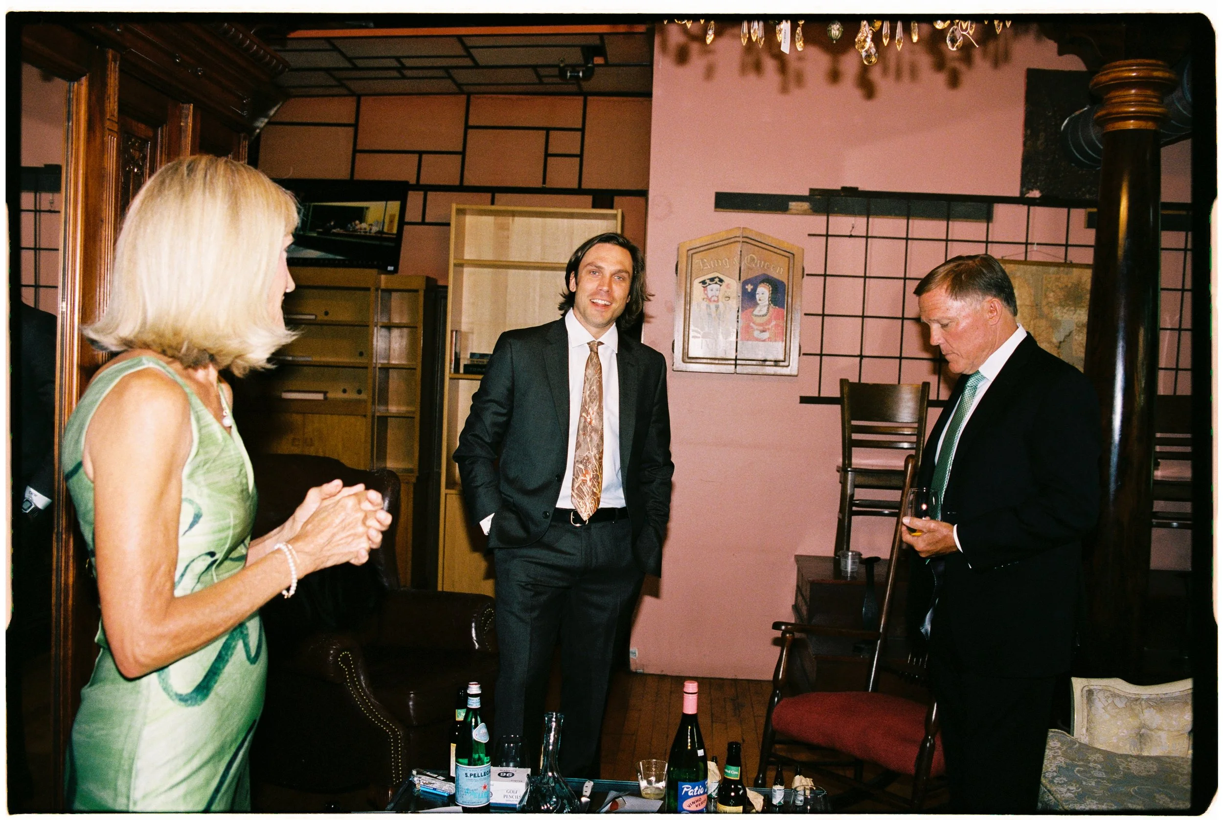 Group of four people gathered in a dimly lit room, with a woman wearing a green dress on the left, a man in a black suit with a patterned tie in the center, and another man in a dark suit with a green tie on the right, holding a drink. The background