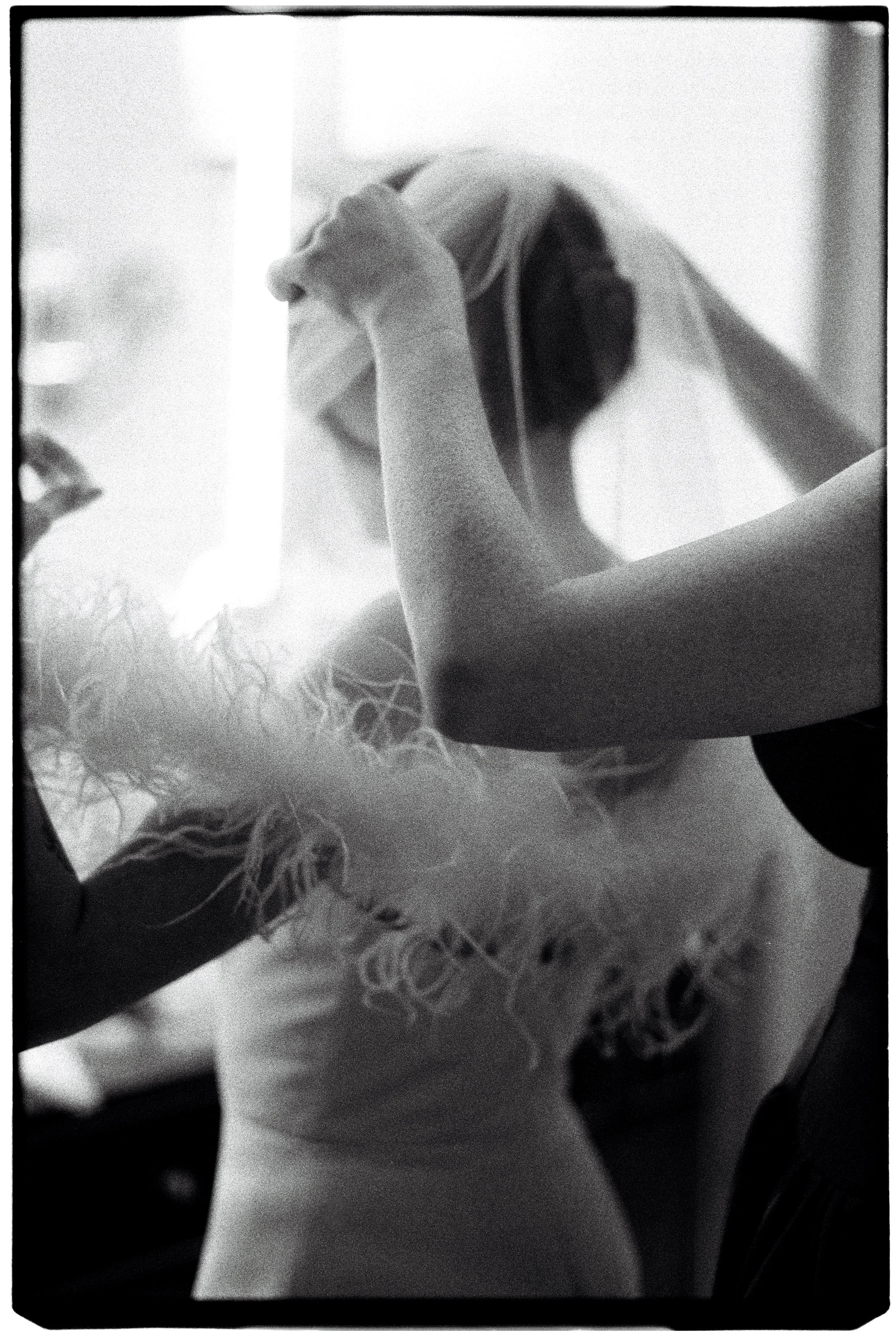 A person with long hair covered by a hood, holding their forehead with one hand, standing in a well-lit indoor space with a lace tablecloth and other objects in the background.