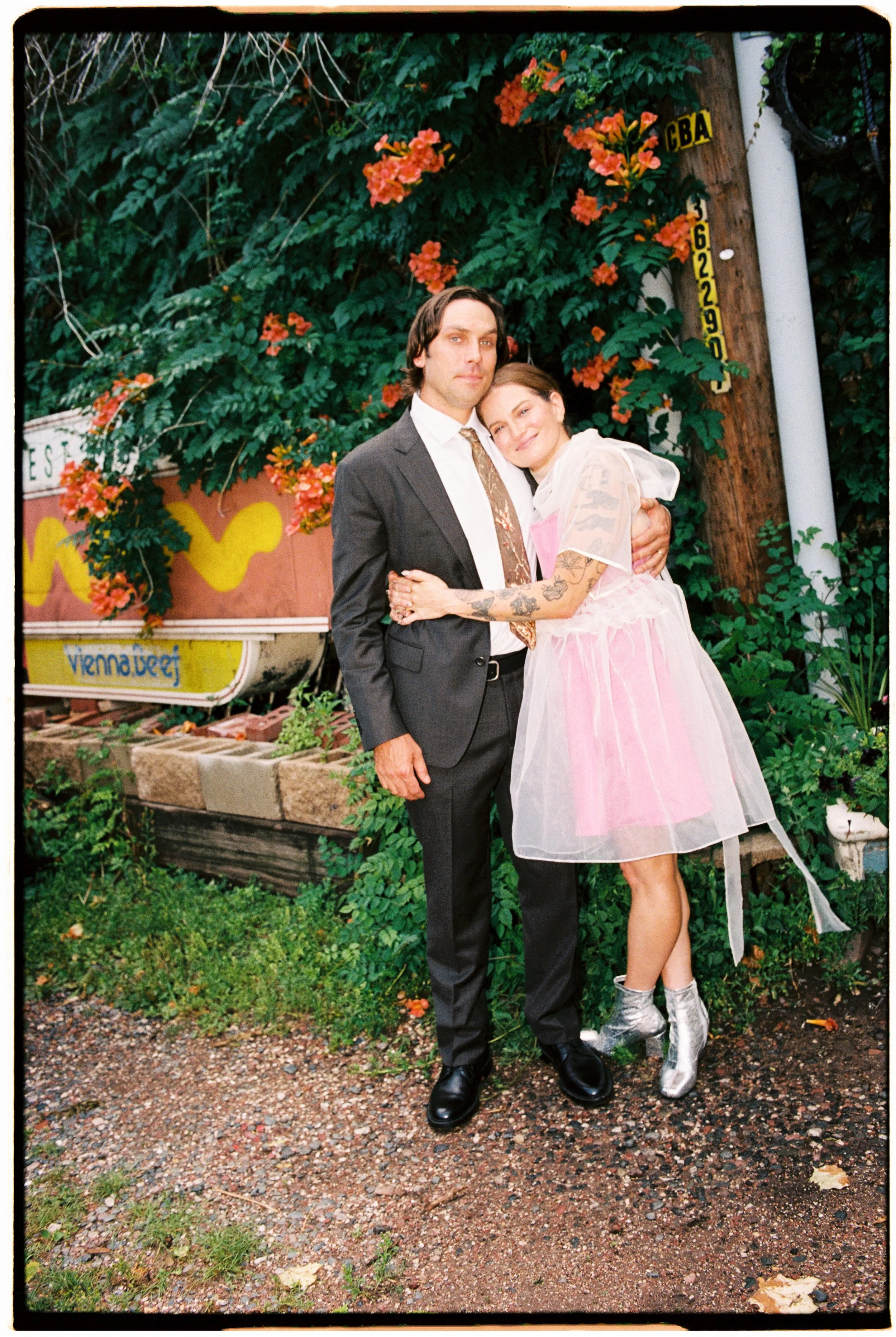 A man and a woman embrace in front of a lush green background with orange flowers, a wooden utility pole, and a yellow sign.
