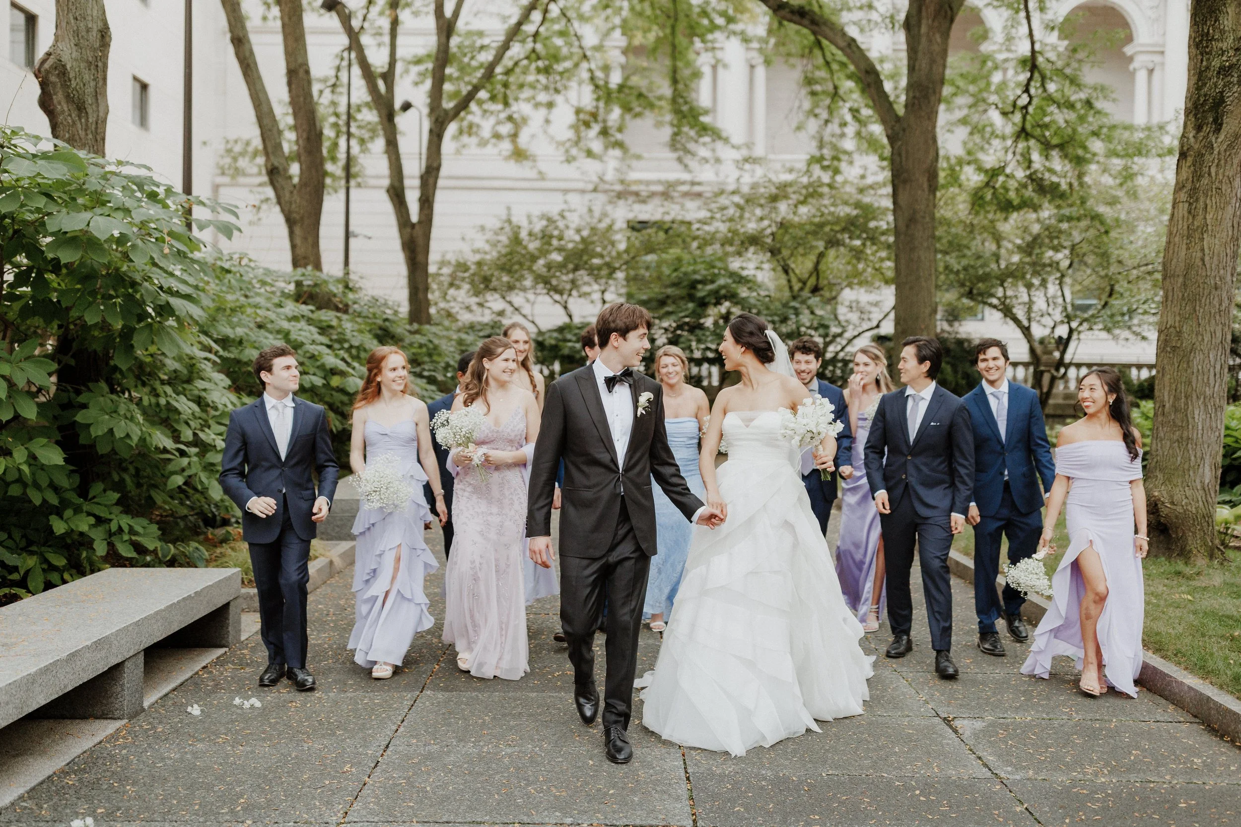 A group of wedding guests, including bridesmaids and groomsmen, walking outdoors in a park with trees and buildings in the background, celebrating a wedding.