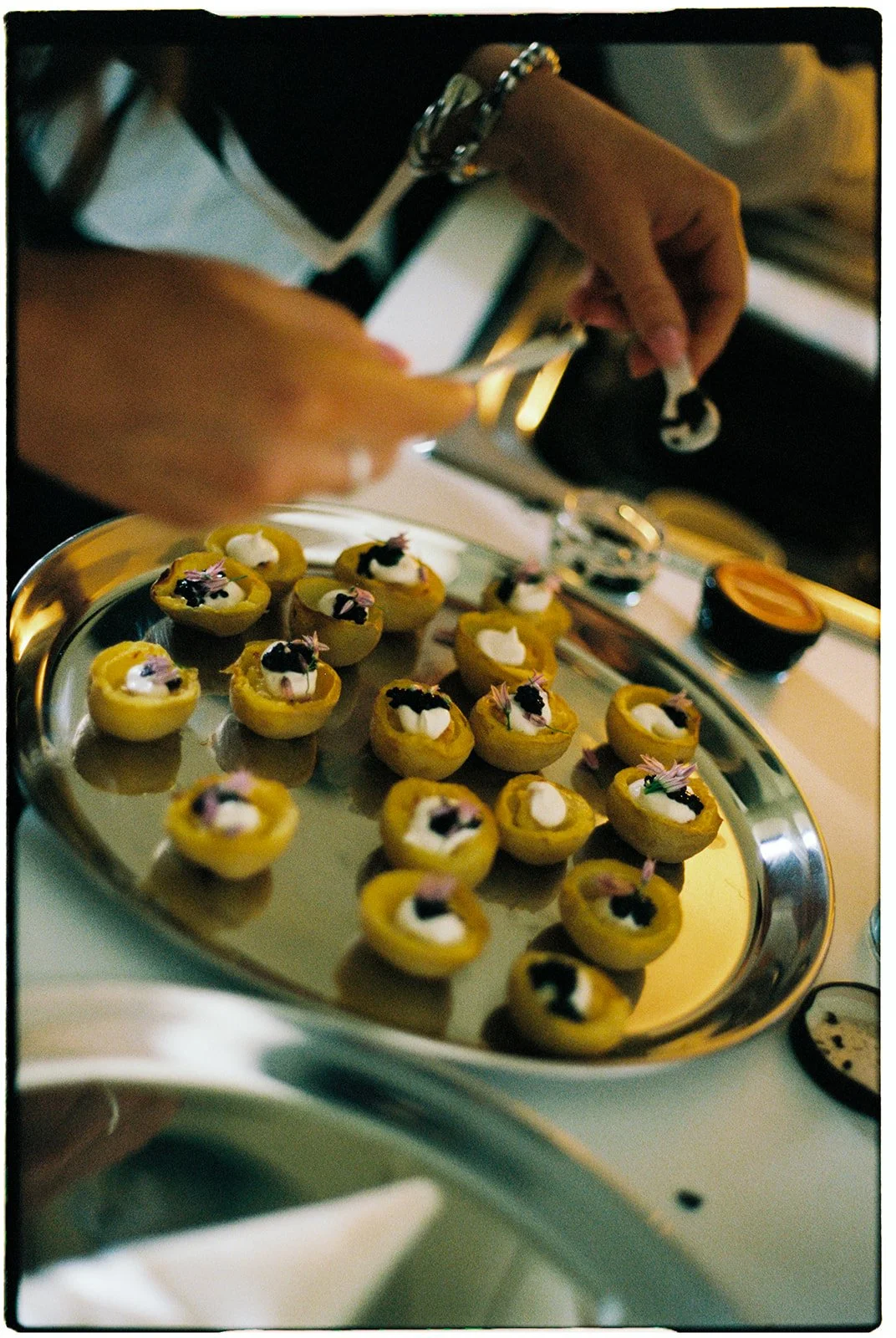 Hands preparing small appetizers or canapés with a silver tray filled with bite-sized food topped with garnishes in a dimly lit setting.