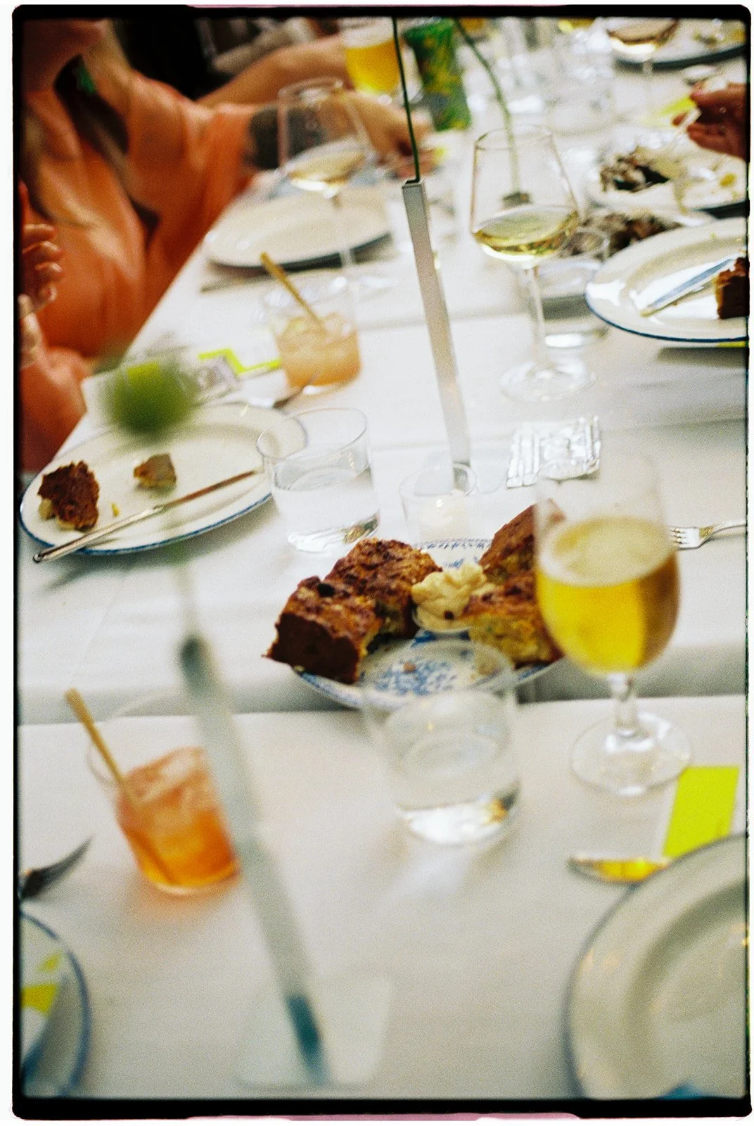 A table set for a meal with plates, glasses of wine, water, and desserts including cake, with people holding drinks around the table.