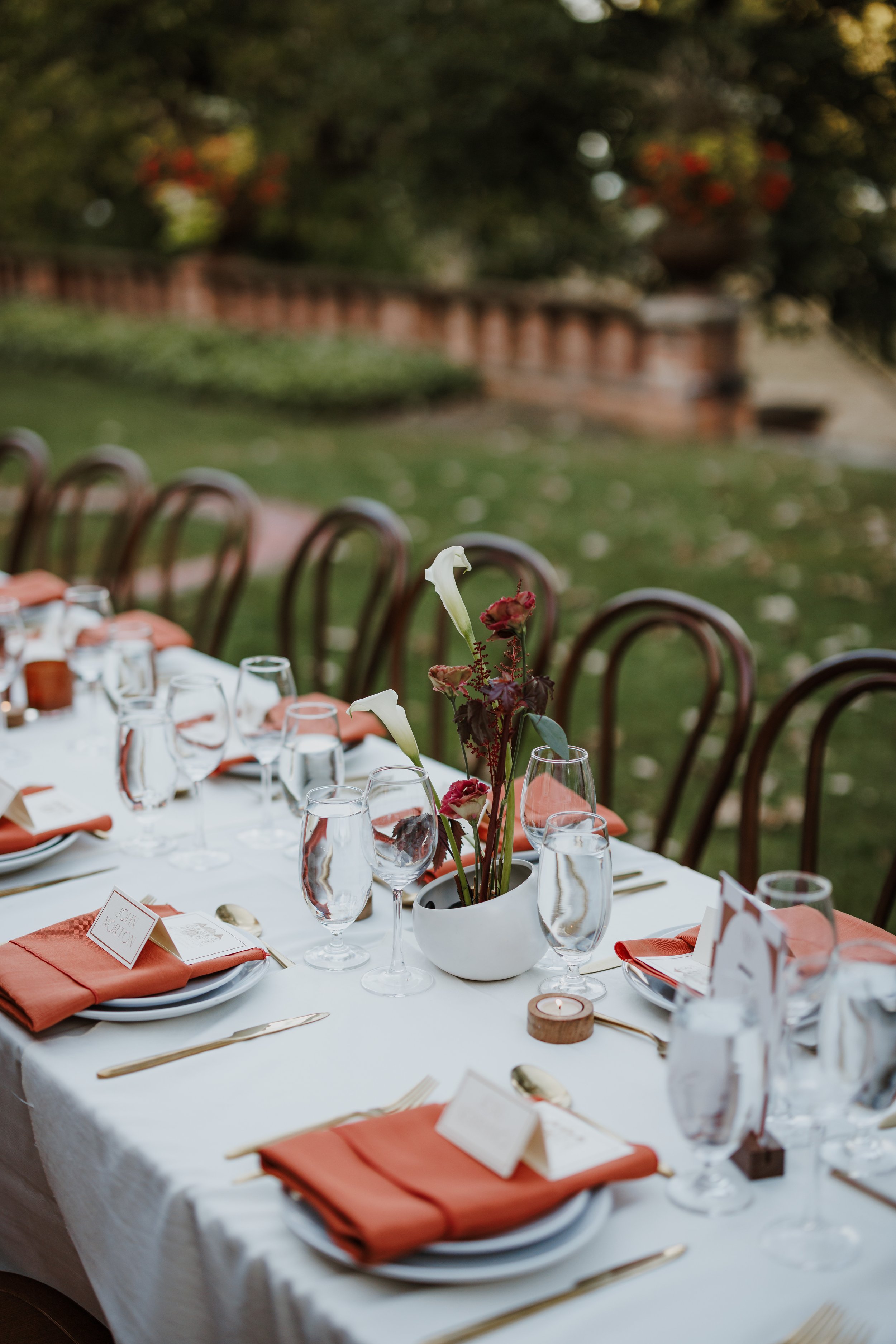 A round dining table set for a formal outdoor event with tableware, glasses, orange napkins, and a floral centerpiece in a white vase. Chairs with wooden backs surround the table, situated in a lush garden with a brick wall and trees in the backgroun
