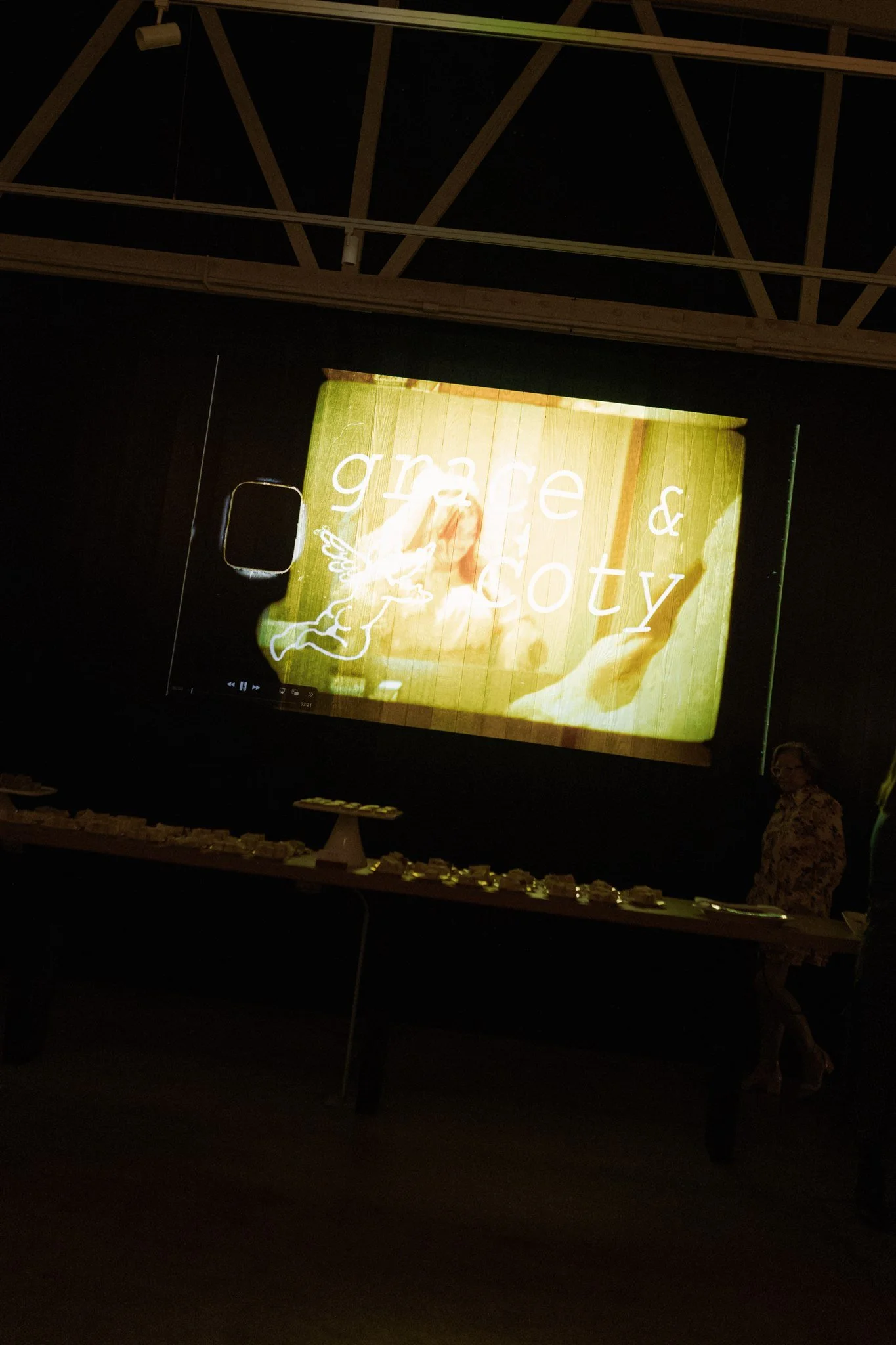A projected slide with the words 'grace & gratitude' on a background of green grass and a dog. A person is standing to the right, partially visible, in a dark room with a wooden ceiling.