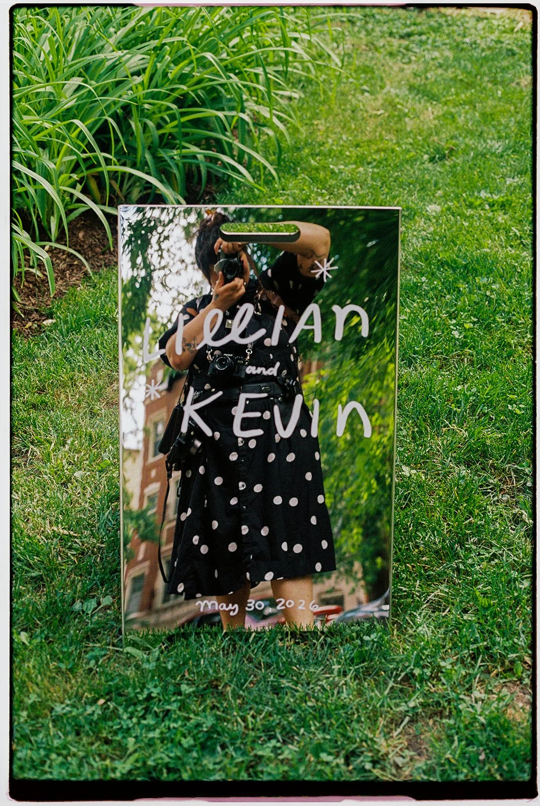 A mirror on the grass reflecting a person taking a photo with a camera. The mirror has handwritten text that says 'Wedding' and 'May 30 2026', along with the names 'Thelian' and 'K.E.V.I.N'.