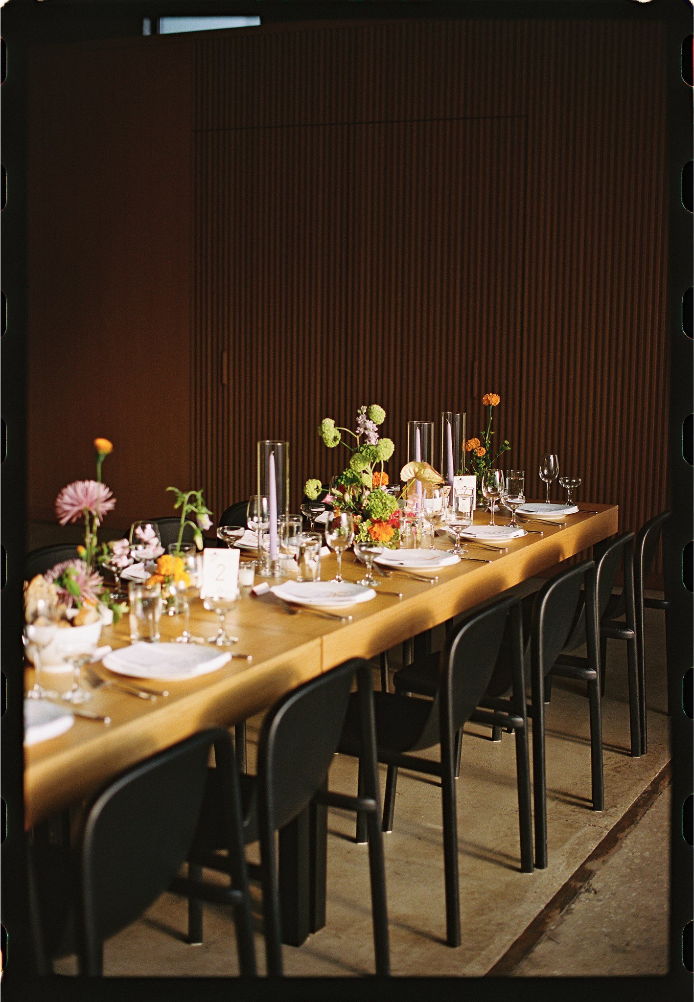 A long wooden dining table decorated with floral centerpieces, candles, and tableware, set for a formal event or dinner in front of a wooden paneled wall.