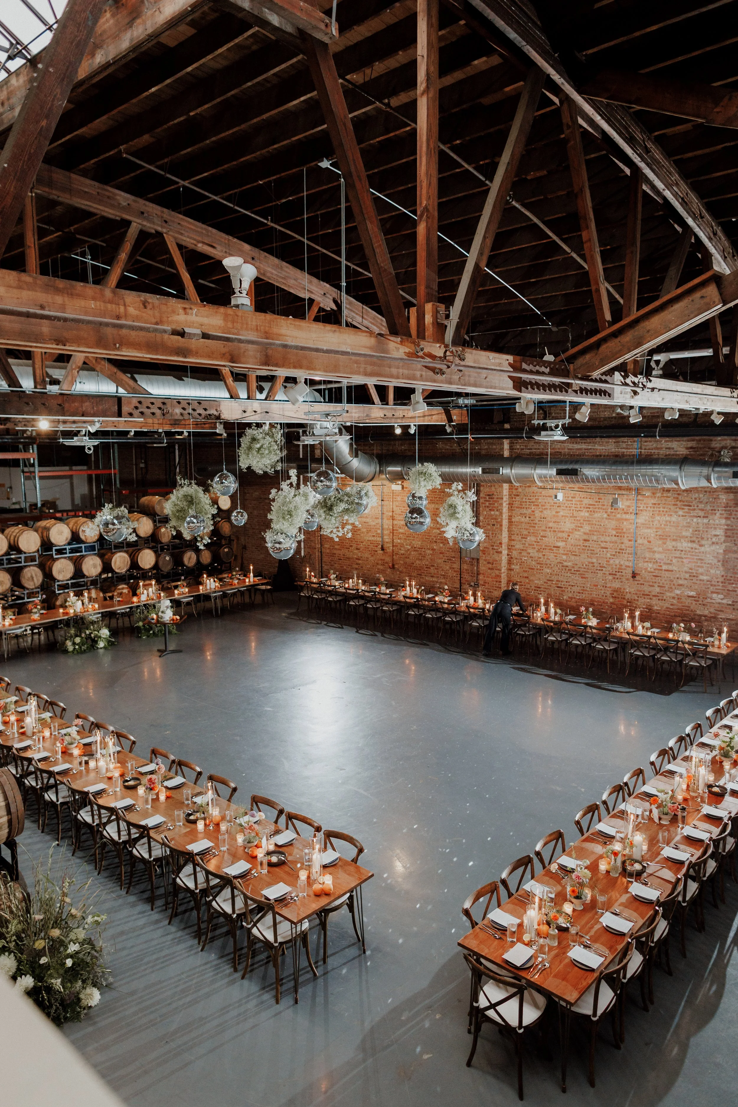 Wedding reception setup with long wooden tables, decorated with candles, flowers, and table settings, in a venue with exposed brick walls and a high wooden ceiling with beams.