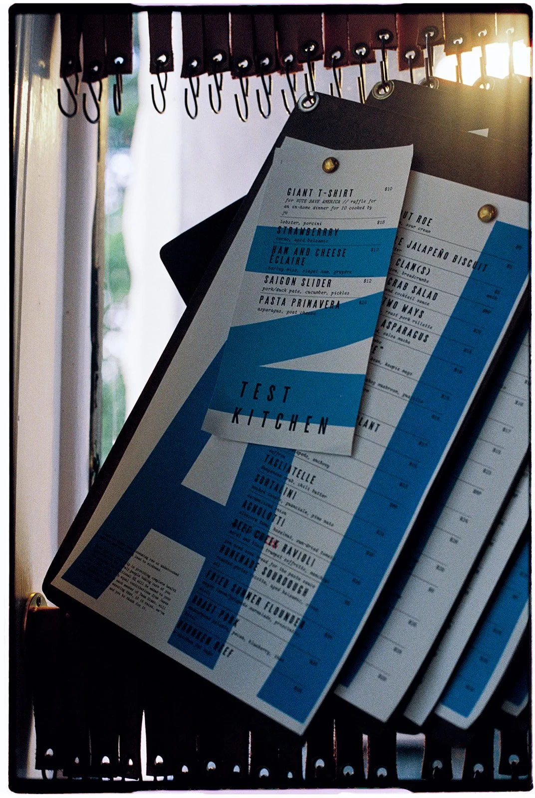 A menu board with a blue and white Test Kitchen design, hanging on hooks from a wooden rack. The menu lists various dishes and prices.