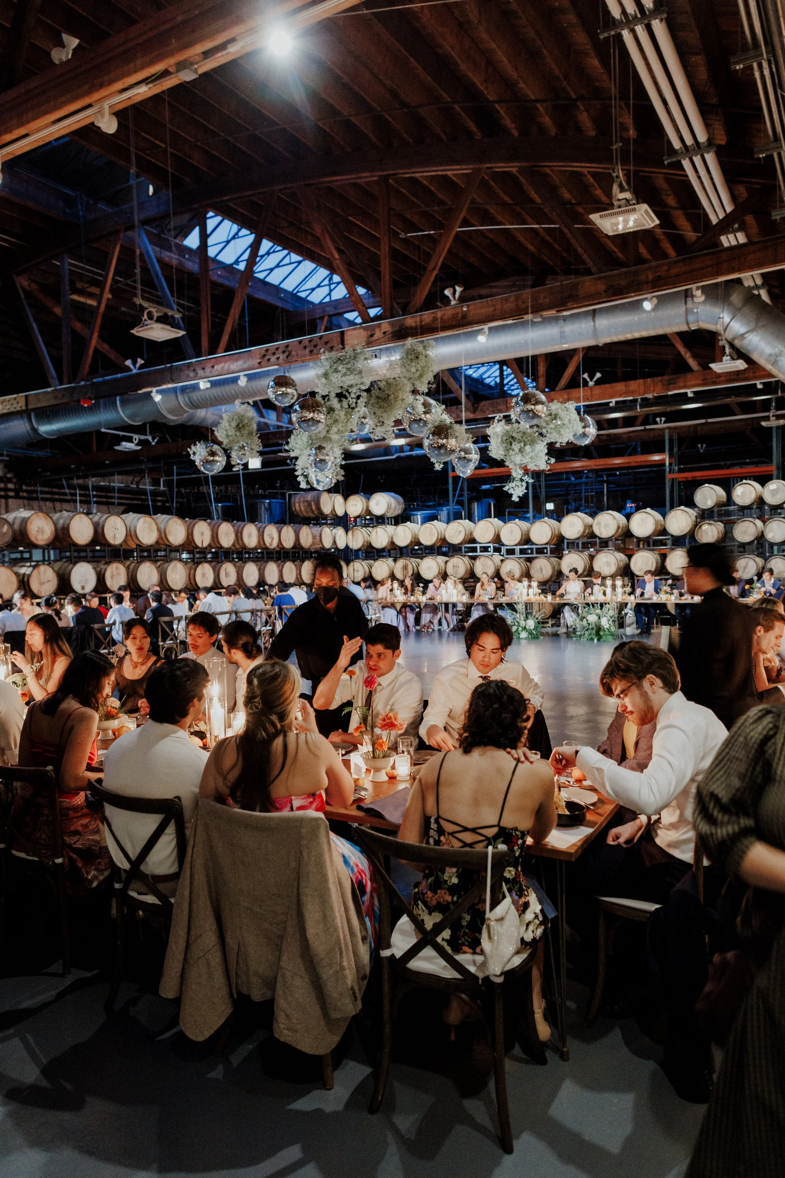 A large indoor dining event with many people seated at tables, decorated with flowers. The ceiling is industrial with wooden beams, ductwork, and hanging floral arrangements and disco balls. The atmosphere is busy and festive.