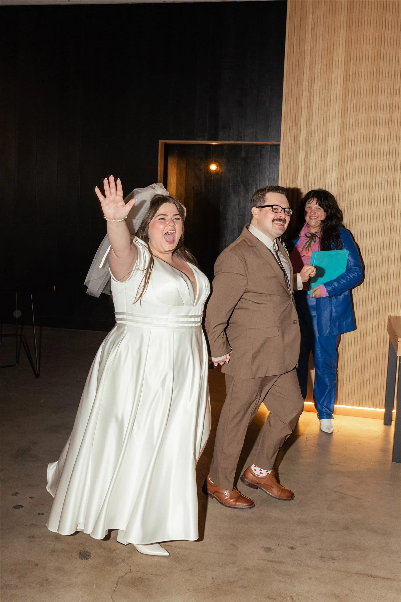 A group of three people celebrating indoors, with the woman on the left wearing a white wedding dress and veil, smiling and waving. The man in the middle is wearing glasses, a tan suit, and brown shoes, smiling. The woman on the right is wearing a co