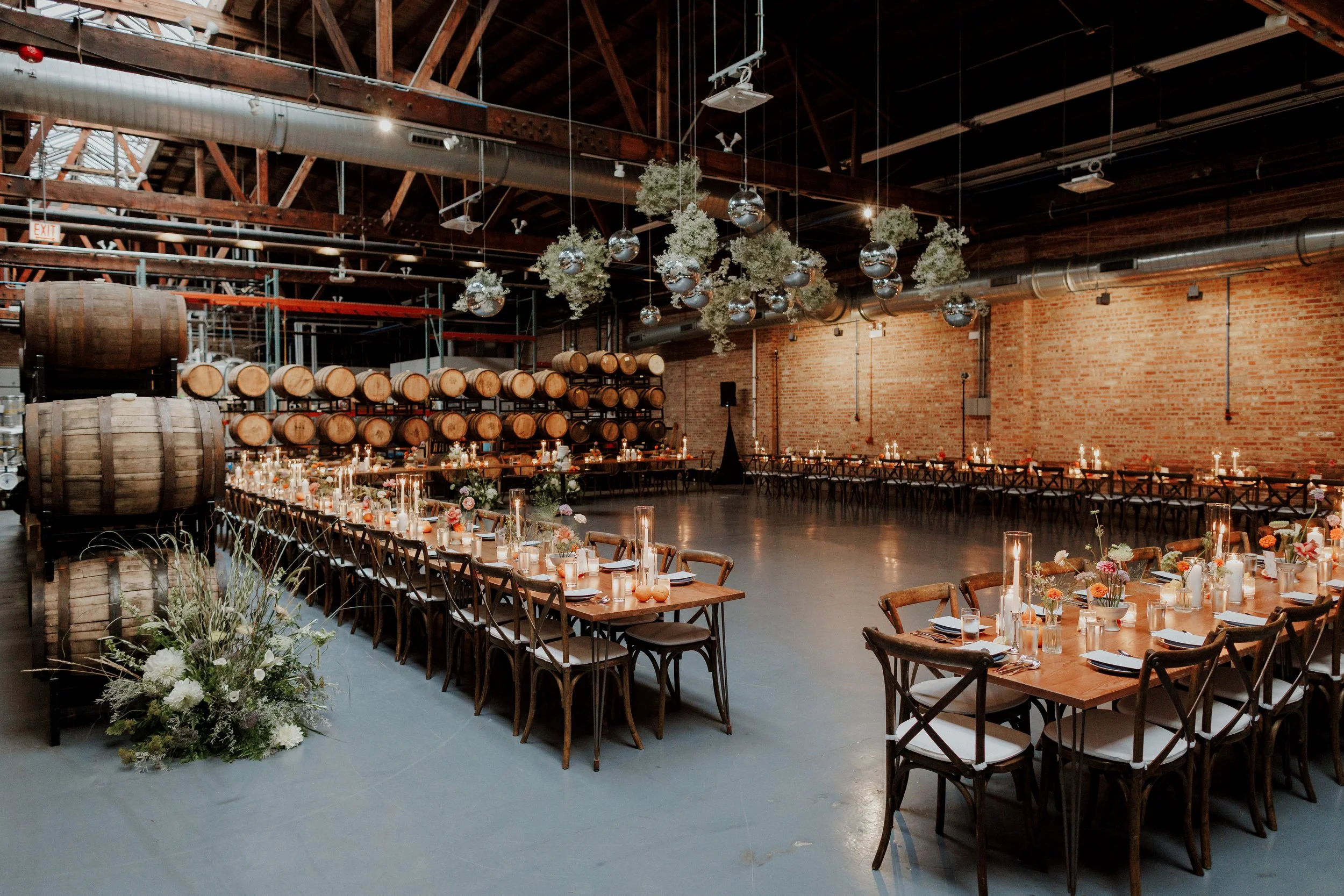 An event space decorated for a celebration with long wooden tables adorned with candles and floral centerpieces. The room features a rustic industrial style with exposed brick walls, wooden beams, and wine barrels stacked on racks. Hanging greenery a