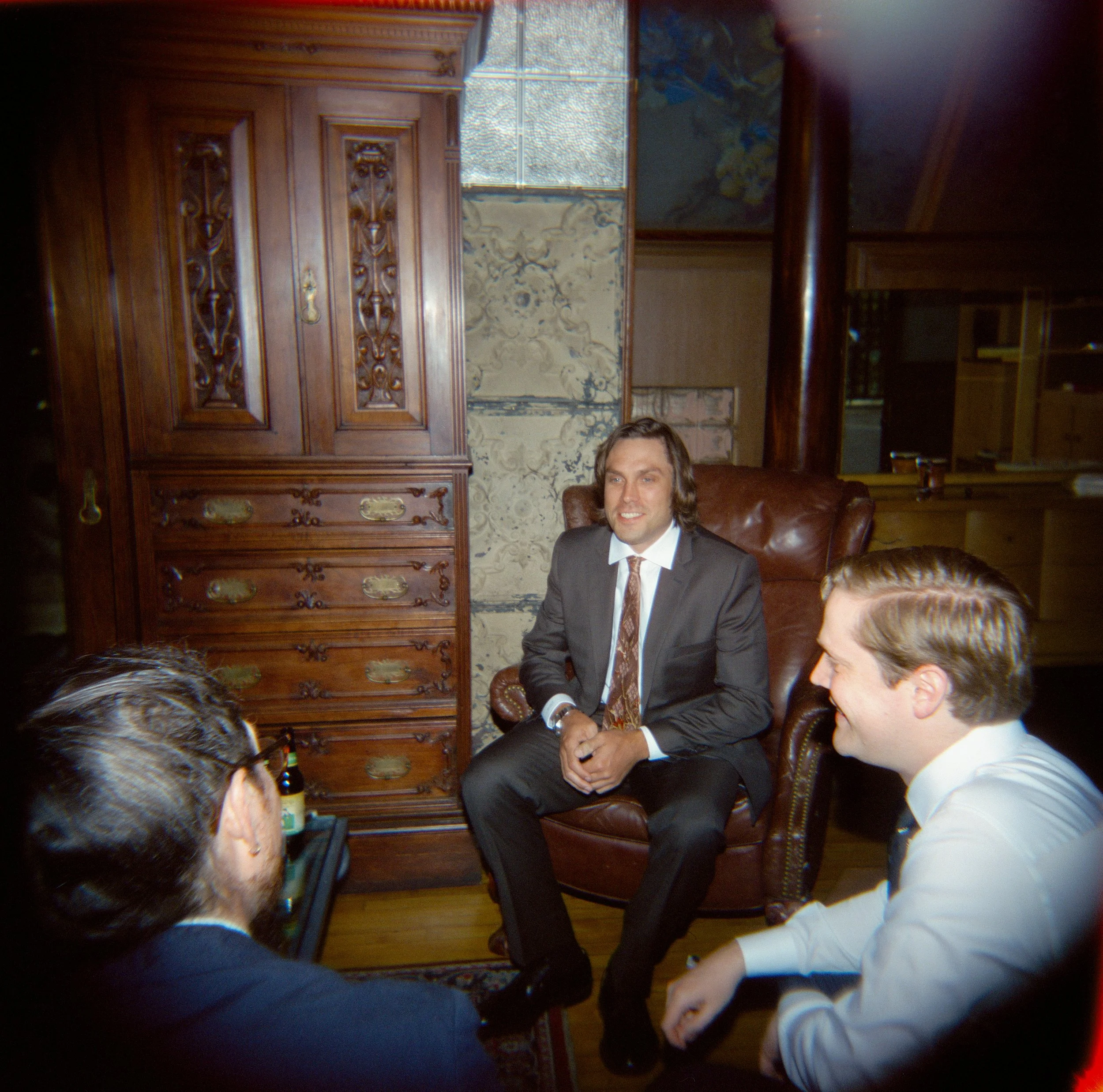 A man in a suit sitting in a leather chair in a room with wooden furniture and decorative stone walls, engaging in a conversation with children.
