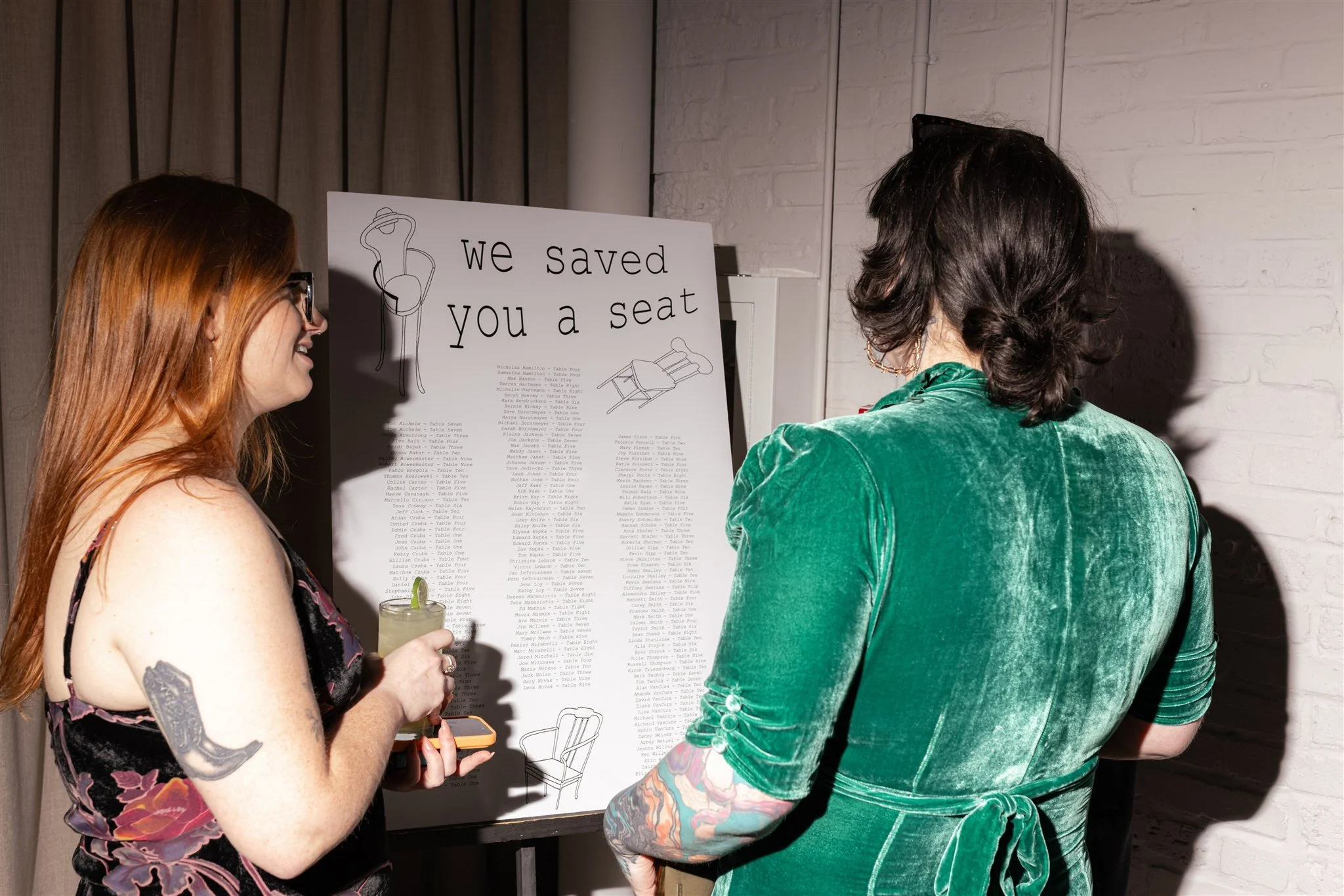 Two women stand in front of a seating chart with the sign, 'we saved you a seat' at an event. One woman has red hair, glasses, and tattoos on her arm, holding a drink and a phone. The other has dark hair, a green velvet jacket, and colorful tattoos o