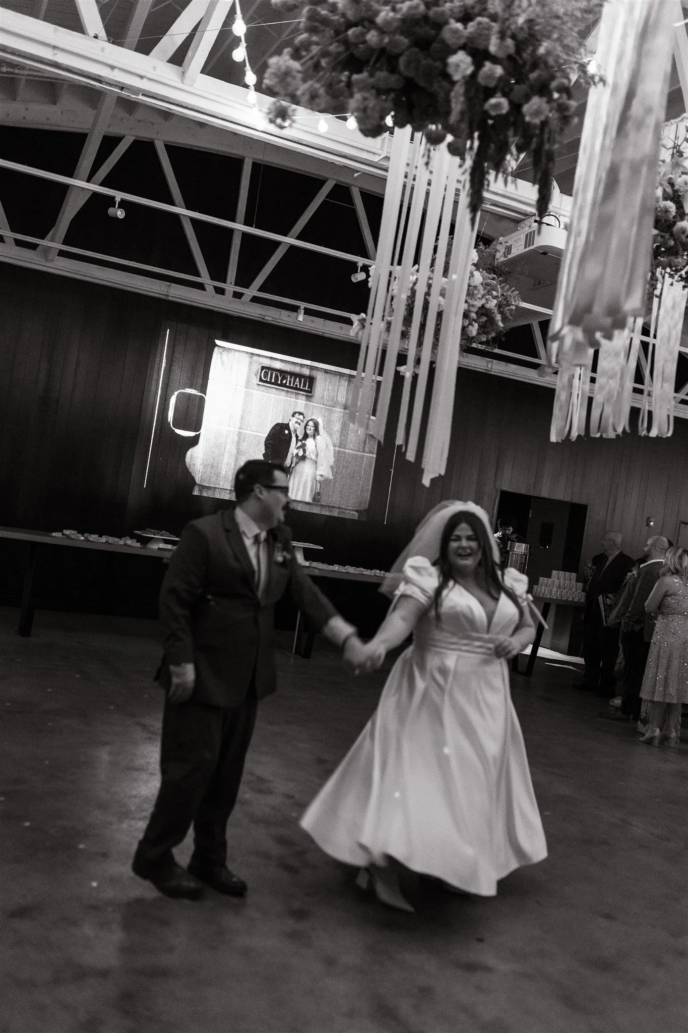 A bride and groom holding hands and dancing at their wedding reception inside a hall with wooden floors and a wooden wall, a large screen showing their photo, and floral decorations hanging from the ceiling.