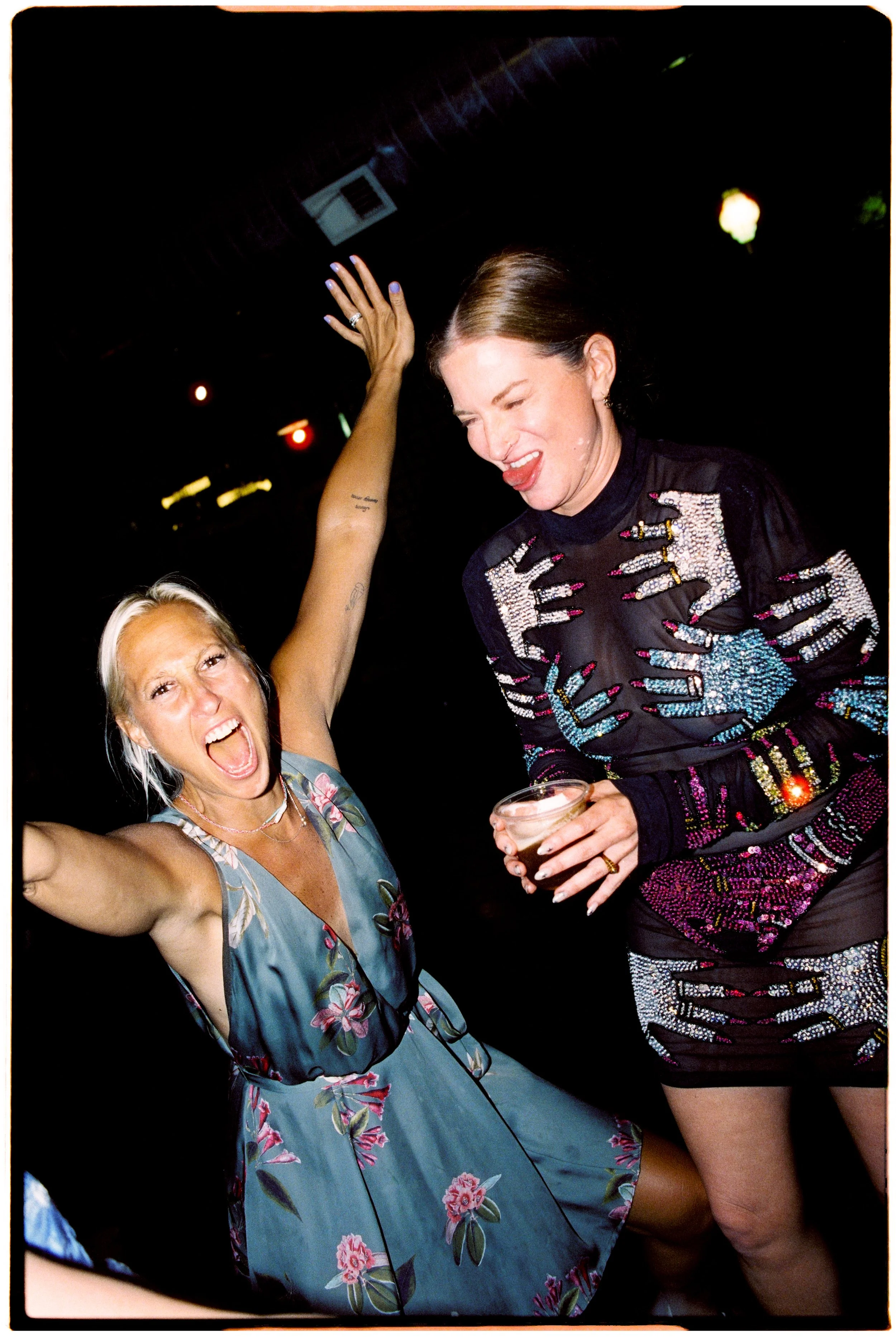 Two women celebrating at a party, one holding a drink, smiling and dancing, with a dark background and colorful lights.