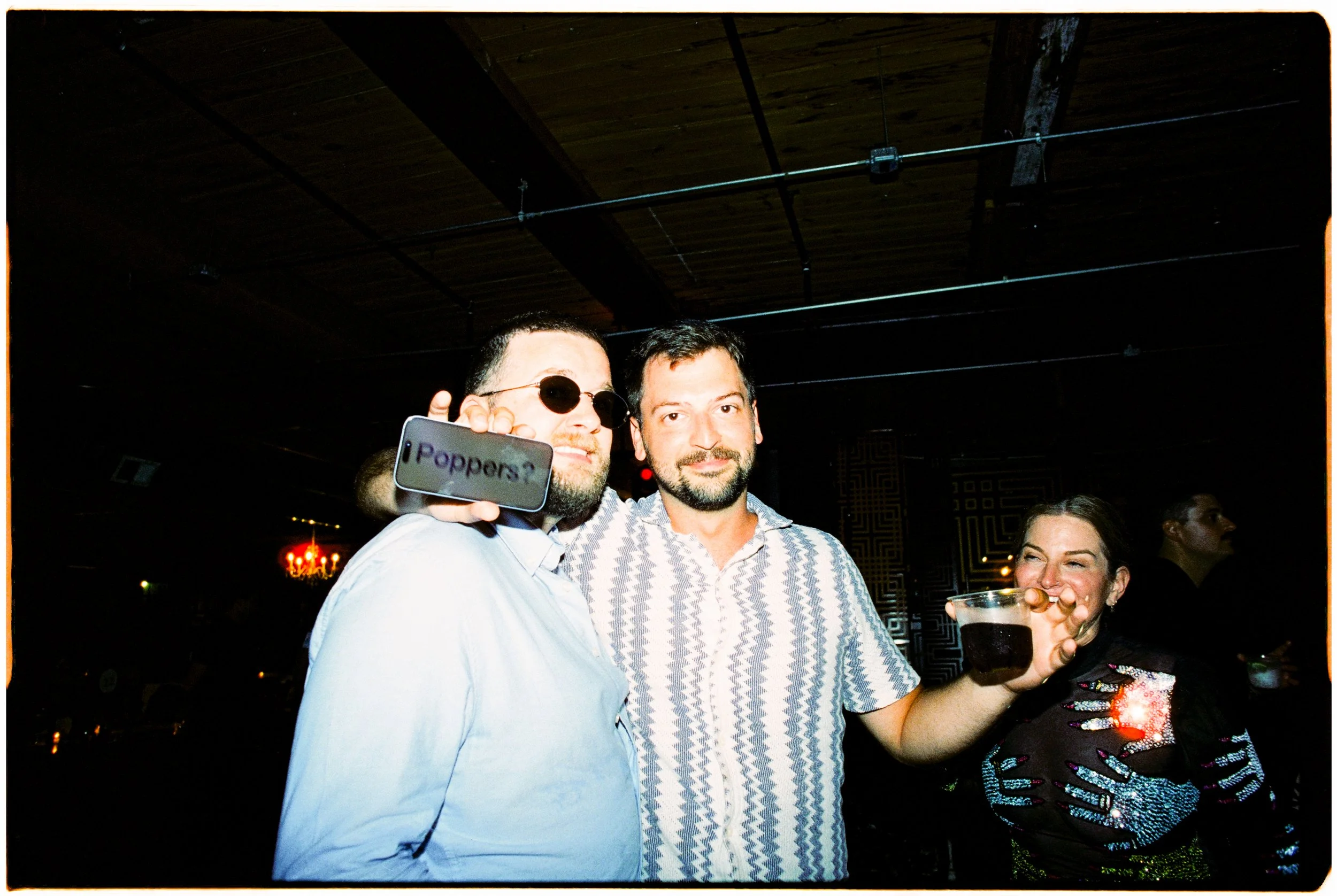 Three people at a social gathering, two men and one woman, holding drinks and smiling, in a dimly lit venue with a dark ceiling and decorative lighting.