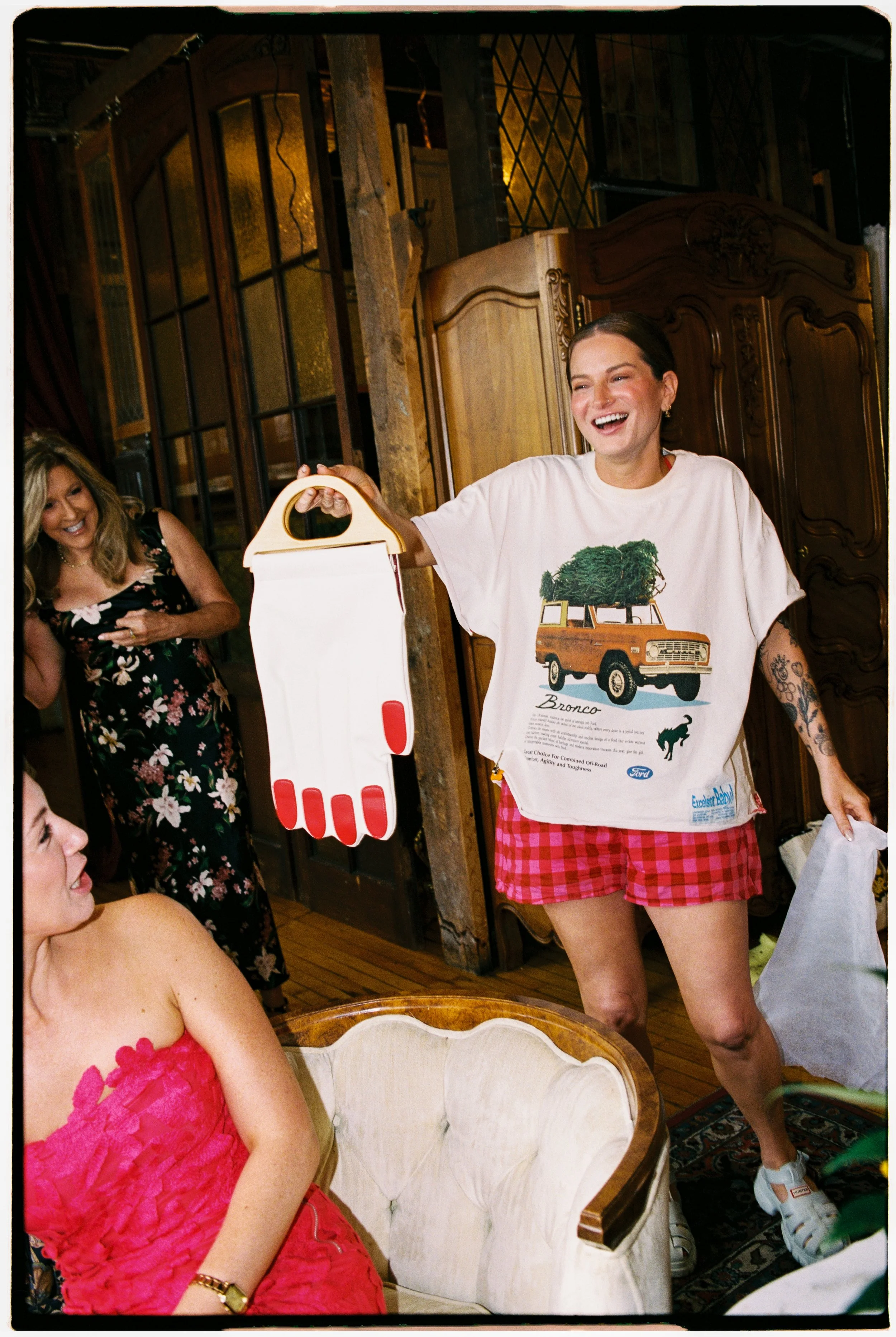 A woman with short dark hair smiling and wearing a graphic t-shirt featuring a vintage SUV with a Christmas tree on top, and red plaid shorts. She is holding a large white and red oven mitt, standing in a cozy wooden room with other women, one in a f