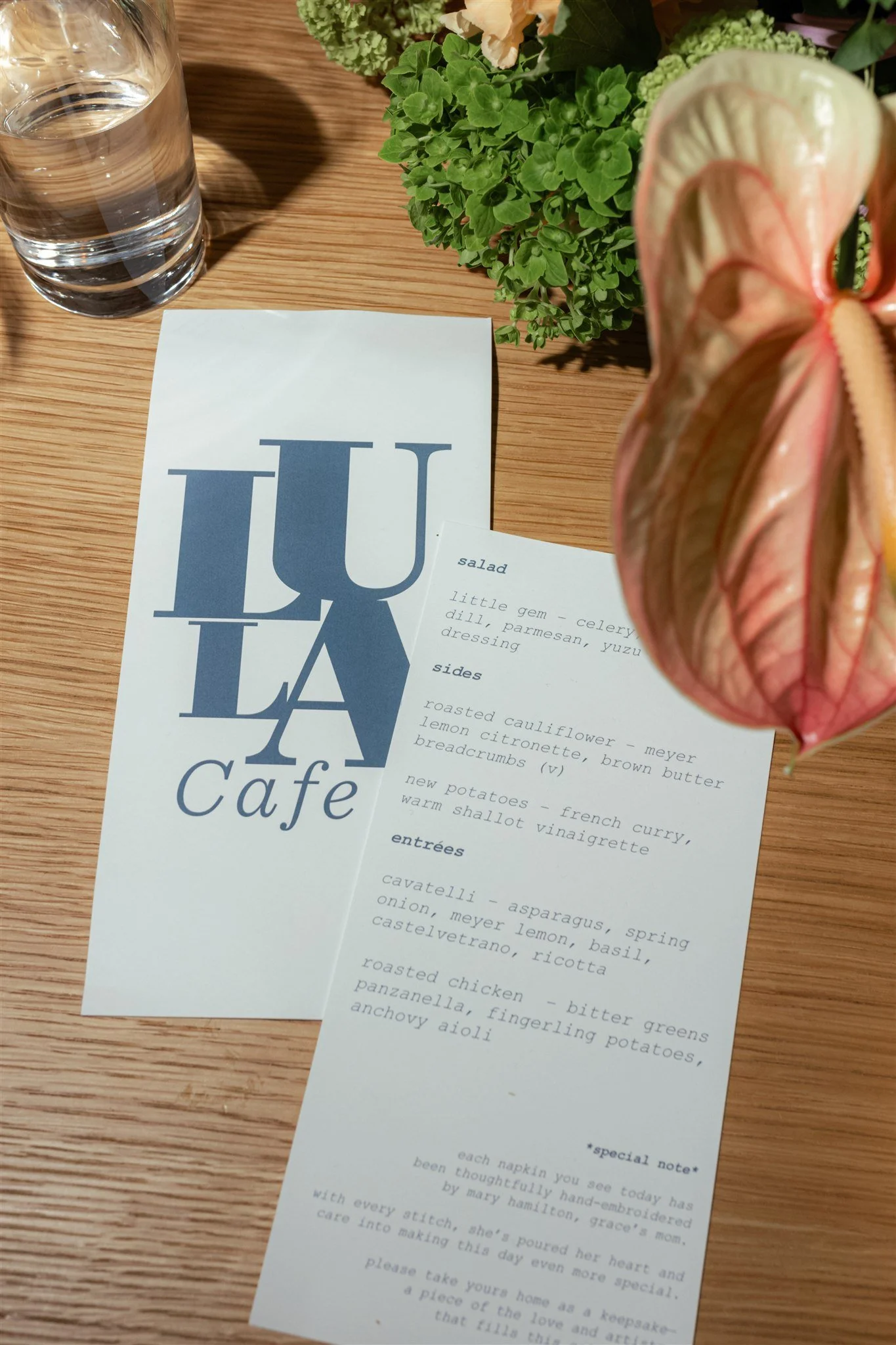 A framed dinner menu from LA Café, placed on a wooden table, with a clear glass of water, some green plants, and a large pink and cream-colored anthurium flower partially blocking the menu. The menu lists various salads, sides, and entrees.