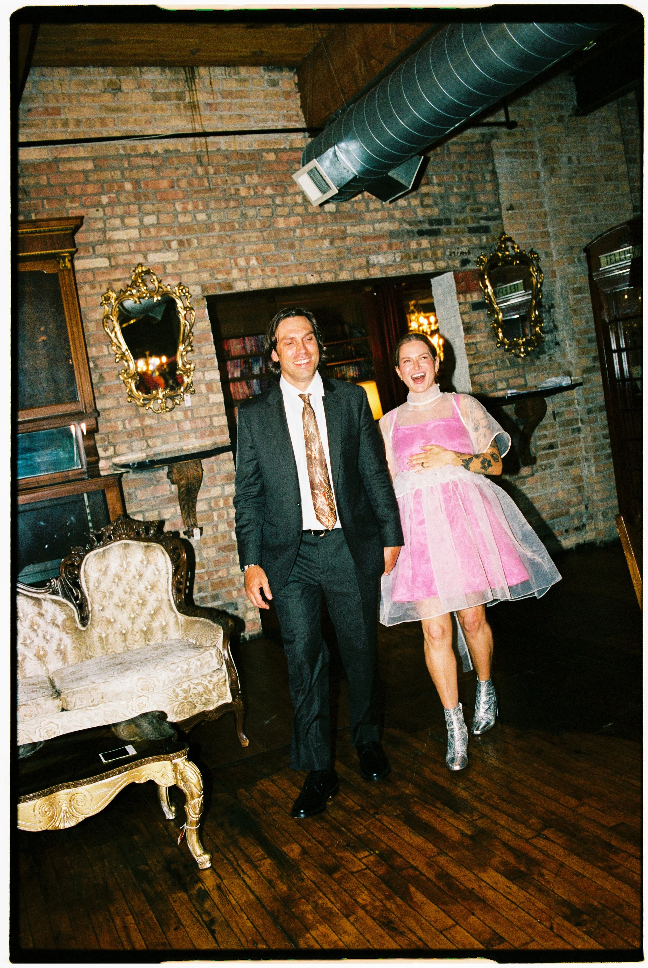 A man in a black suit with a patterned tie and a woman in a pink dress with lace sleeves and silver boots are laughing and smiling inside a room with a brick wall, vintage mirrors with gold frames, and old-fashioned furniture.